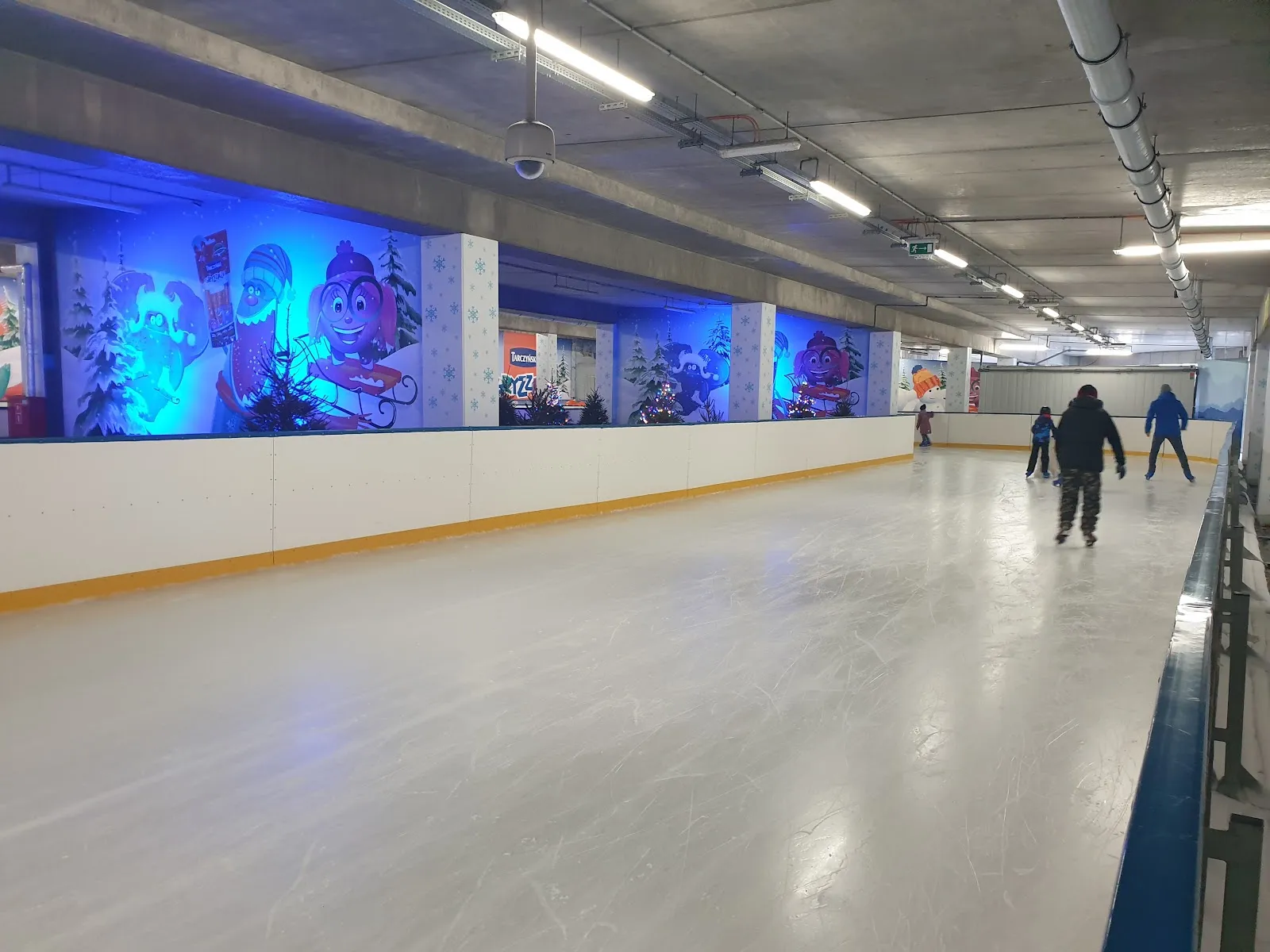 Długa owalna tafla pod stropem parkingu z niebieskimi bandami ozdobionymi śnieżynkami i kilkoma łyżwiarzami w oddali. — Lodowisko Tarczyński Arena