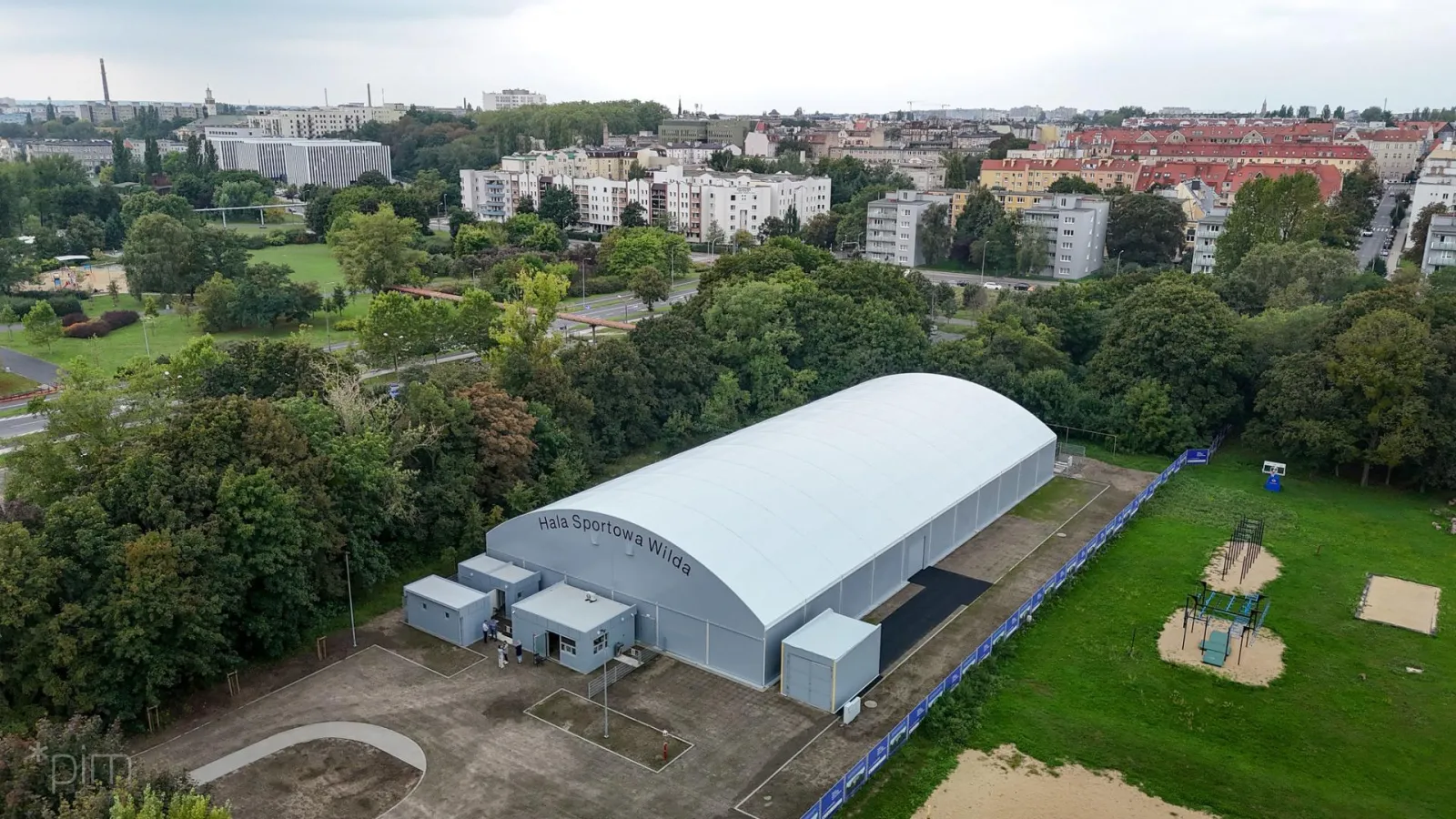 Widok z lotu ptaka na białą łukową Halę Sportową Wilda w otoczeniu parku; w hali działa sezonowe lodowisko — Lodowisko w Hali Sportowej Wilda