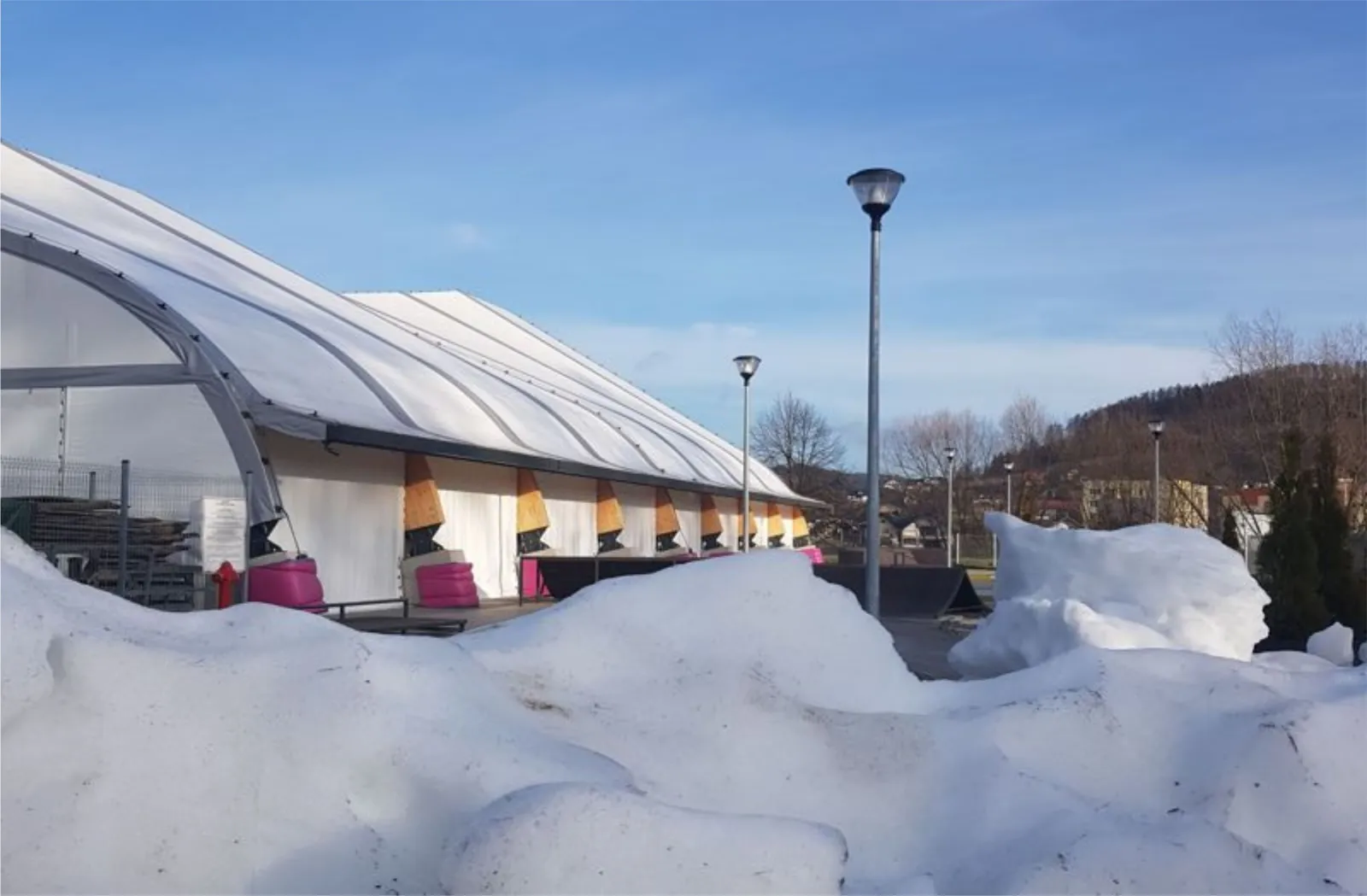 The white arched tent hall of the Muszyna rink on a sunny winter day, with timber and pink panels along its side, lamp posts and a skate-ramp beyond, and snow banked in the foreground. — Lodowisko Zapopradzie Muszyna