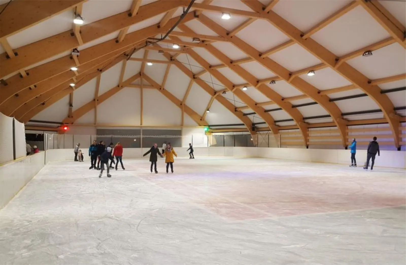 Skaters, some holding hands, on the boarded ice inside the timber-framed roofed hall of the Muszyna rink, with laminated wooden arches and floodlights overhead. — Lodowisko Zapopradzie Muszyna