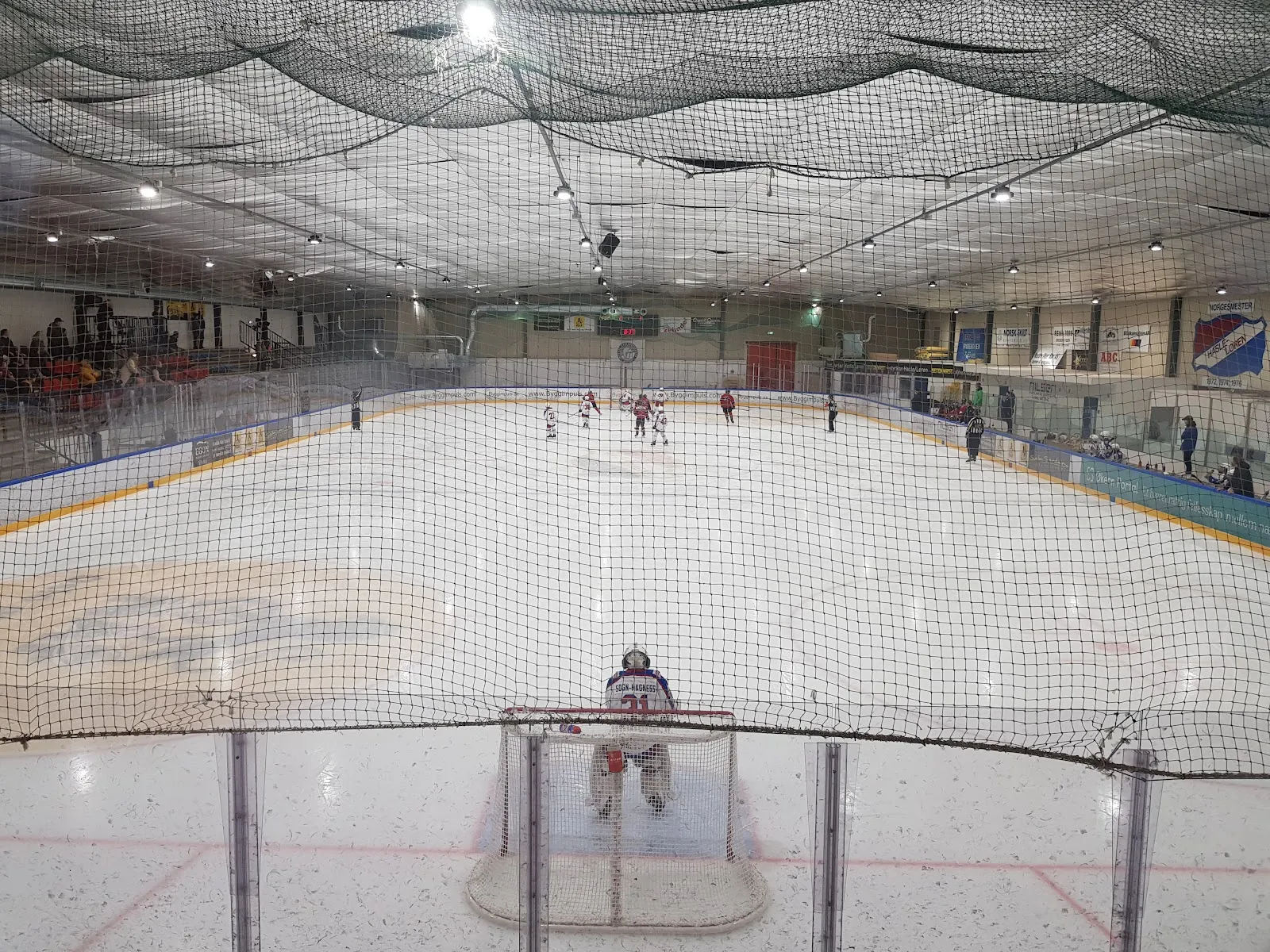 Wide view across the indoor rink from the end boards, with a game in progress and the far goal in view. — Løren ishall