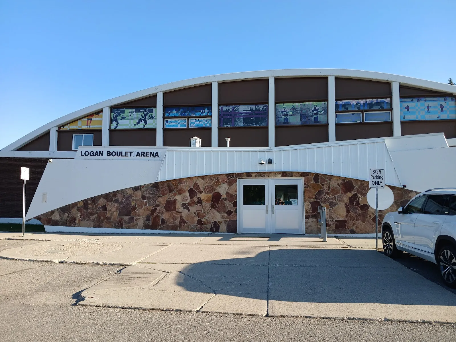 El frente de techo curvo de Logan Boulet Arena en Lethbridge, con el nombre de la arena sobre un muro de piedra de campo y las puertas de entrada — Logan Boulet Arena