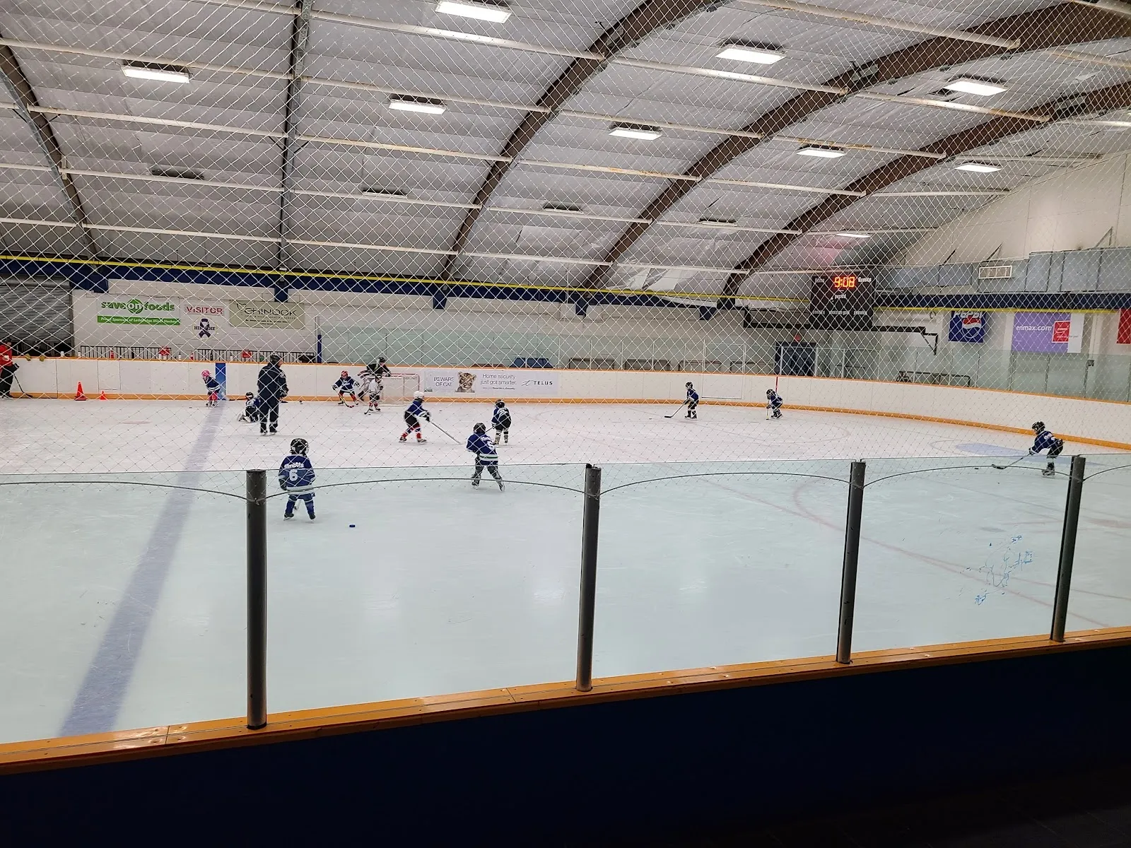 Un partido visto a través de una pista amplia desde detrás del vidrio, con jugadores repartidos por el hielo bajo un largo techo arqueado con un barandal verde y blanco. — Logan Boulet Arena