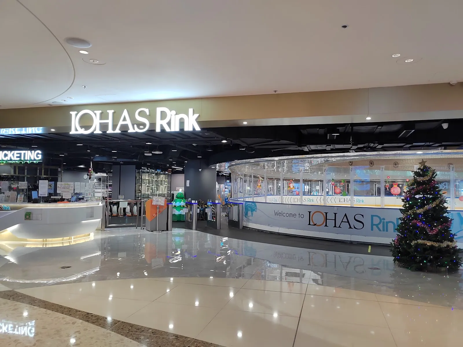 康城內入口與閘口上方亮著的 LOHAS Rink 招牌，旁邊是溜冰場圍板，右側有一棵聖誕樹。 — LOHAS Rink