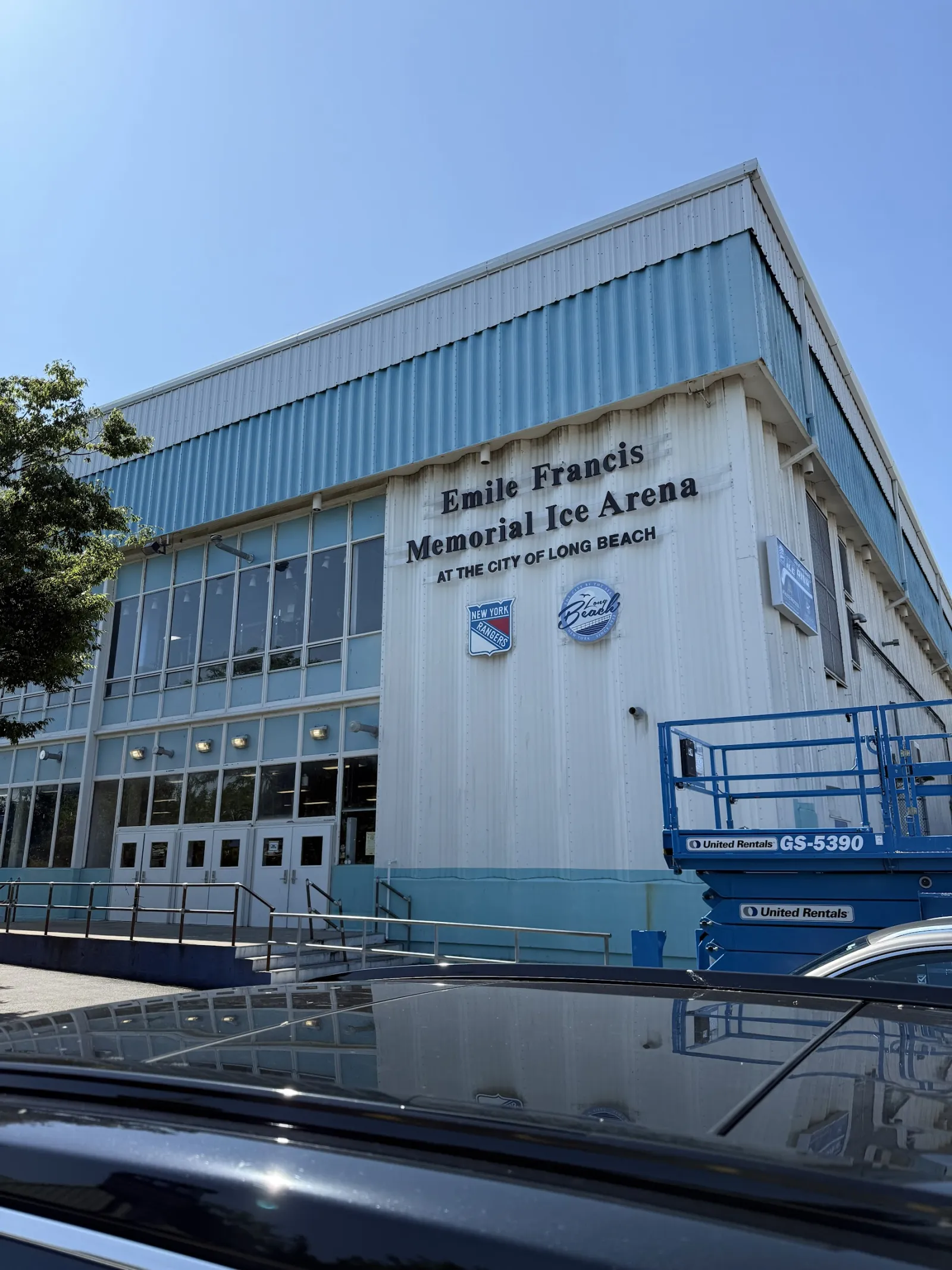 El edificio azul pálido rotulado Emile Francis Memorial Ice Arena at the City of Long Beach, con logotipos de los Rangers y los Islanders junto a la entrada de vidrio. — City of Long Beach Ice Arena