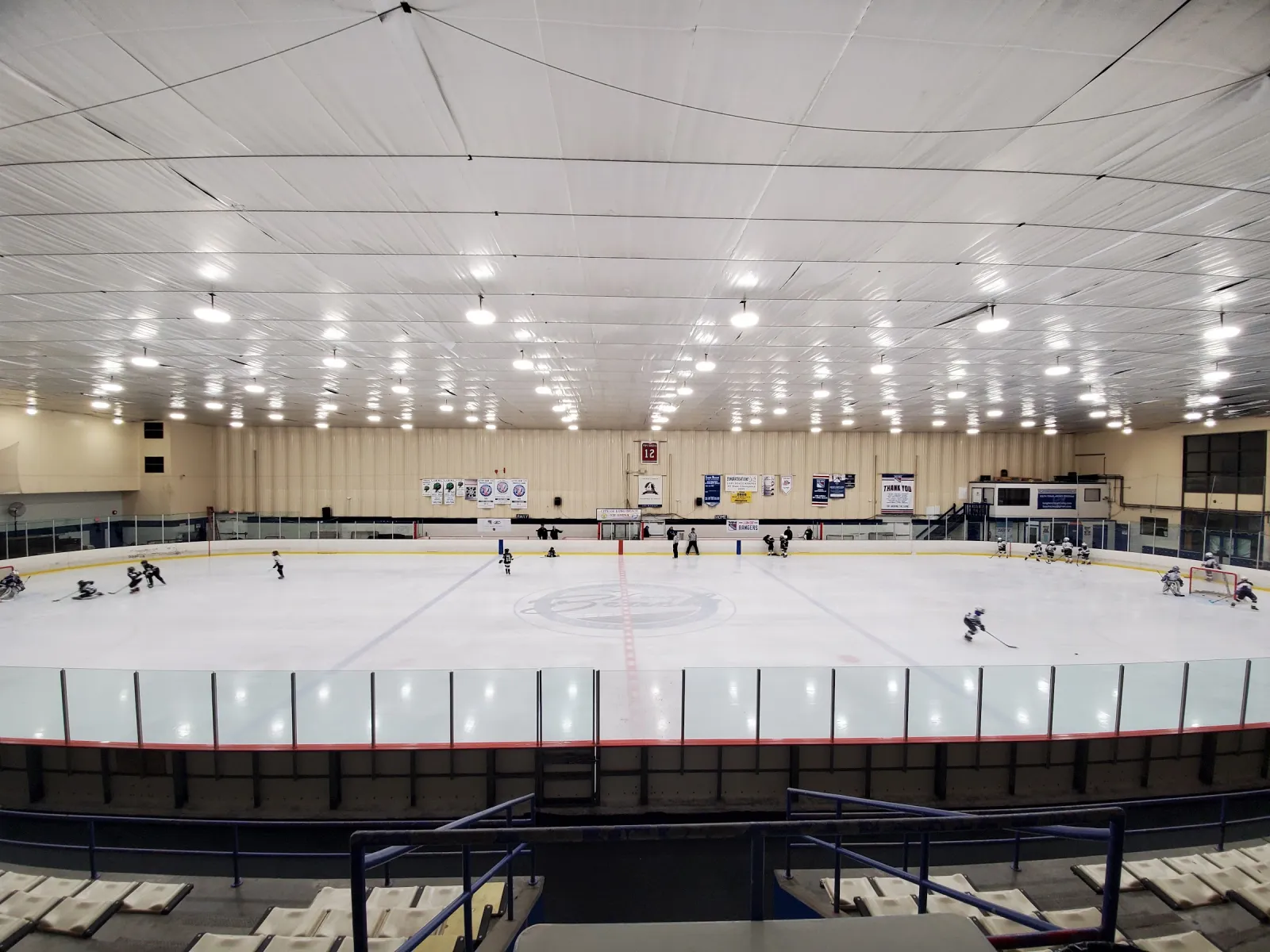 Todo el hielo visto desde lo alto de las gradas, con una sesión de hockey bajo hileras de luces y mantas a lo largo de la pared del fondo. — City of Long Beach Ice Arena