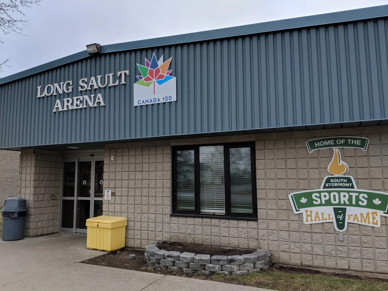 The Long Sault Arena entrance, a blue-clad front with the arena name in large letters, a Canada 150 mark and the South Stormont Sports Hall of Fame sign on the block wall. — Long Sault Arena