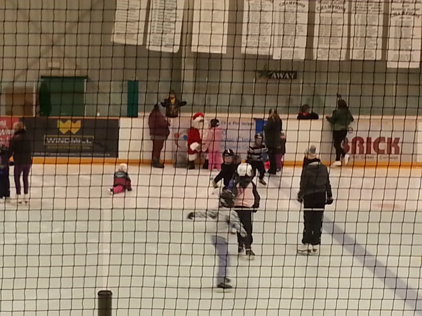 A public skating session at the Long Sault Arena seen through the safety netting, with skaters of all ages across the ice and banners hanging above the boards. — Long Sault Arena