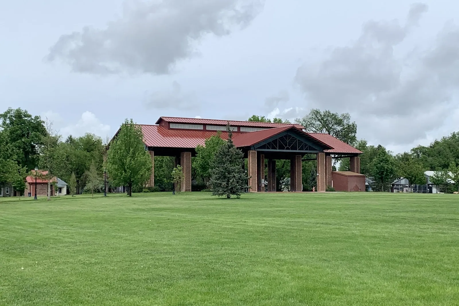 El pabellón abierto de madera y acero sobre la pista de Longmont, un edificio de techo rojo entre árboles al otro lado de un jardín, bajo un cielo nublado. — Longmont Ice Pavilion