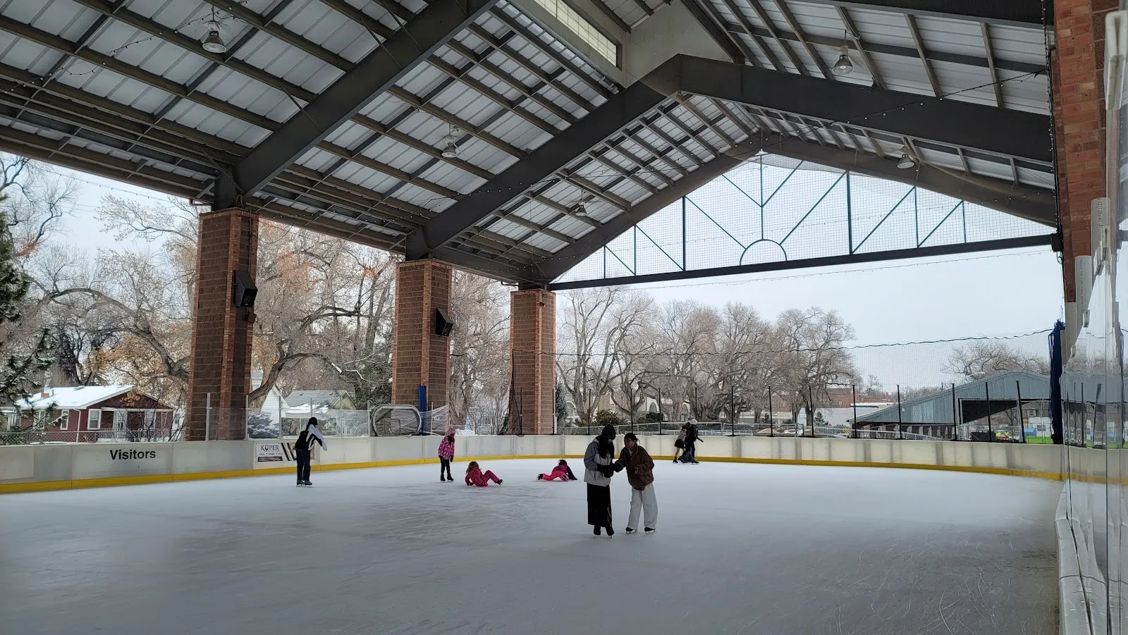 Patinaje público bajo el techo de acero de lados abiertos de Longmont Ice Pavilion, sobre pilares de ladrillo, con árboles sin hojas y casas más allá de las vallas — Longmont Ice Pavilion