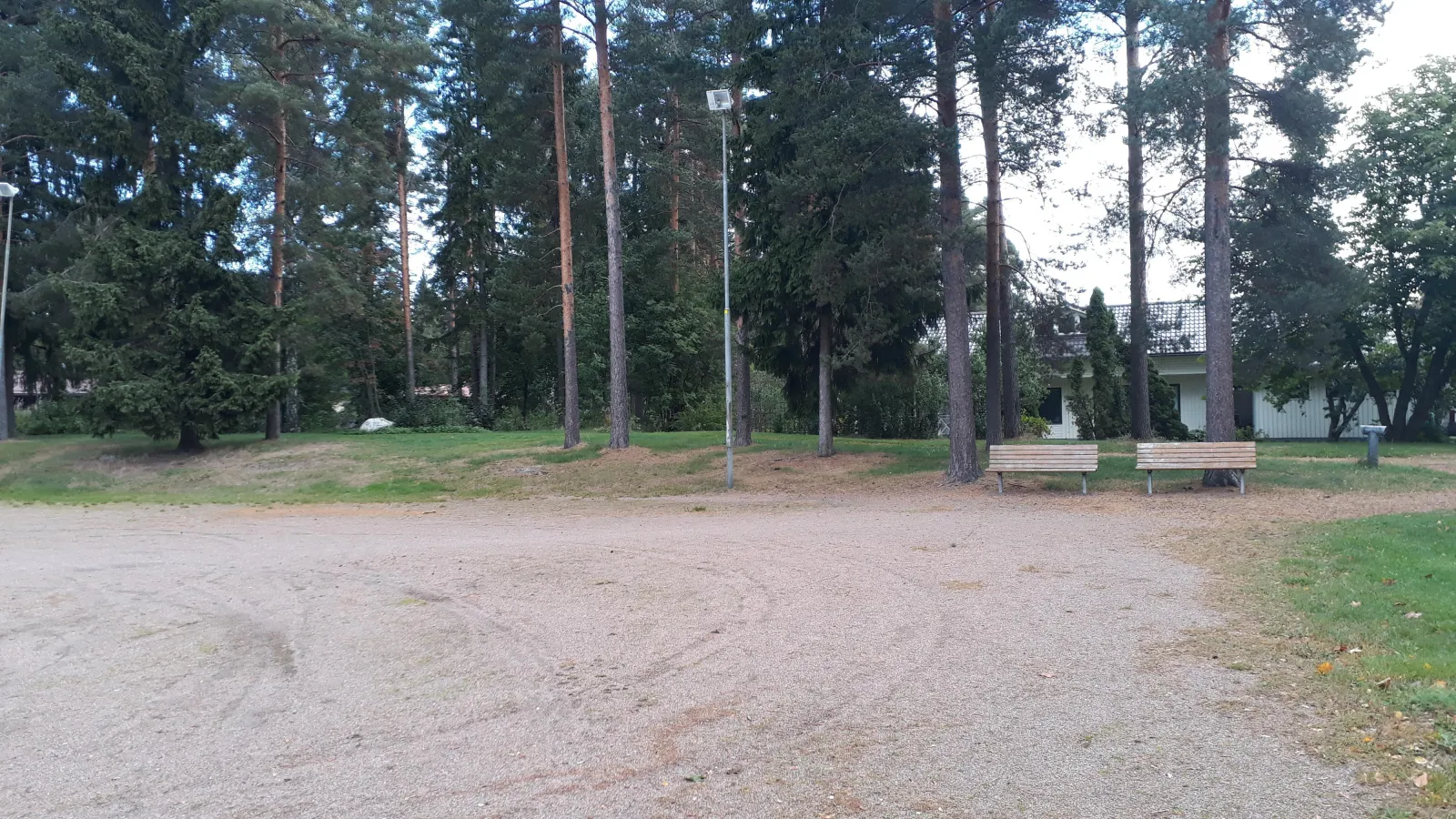 The sand field at Lönnrotinpuisto in Kevätkumpu, which the city freezes for skating each winter when the weather allows, seen in summer with two benches and tall pines behind. — Lönnrotinpuiston luistelukenttä