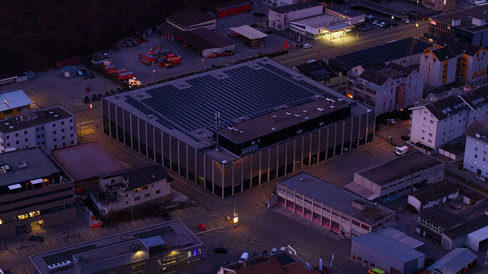 Luftaufnahme der Lonza Arena in Visp in der Dämmerung, die rechteckige Halle mit ihrem Solardach zwischen den Gebäuden der Stadt – Lonza Arena
