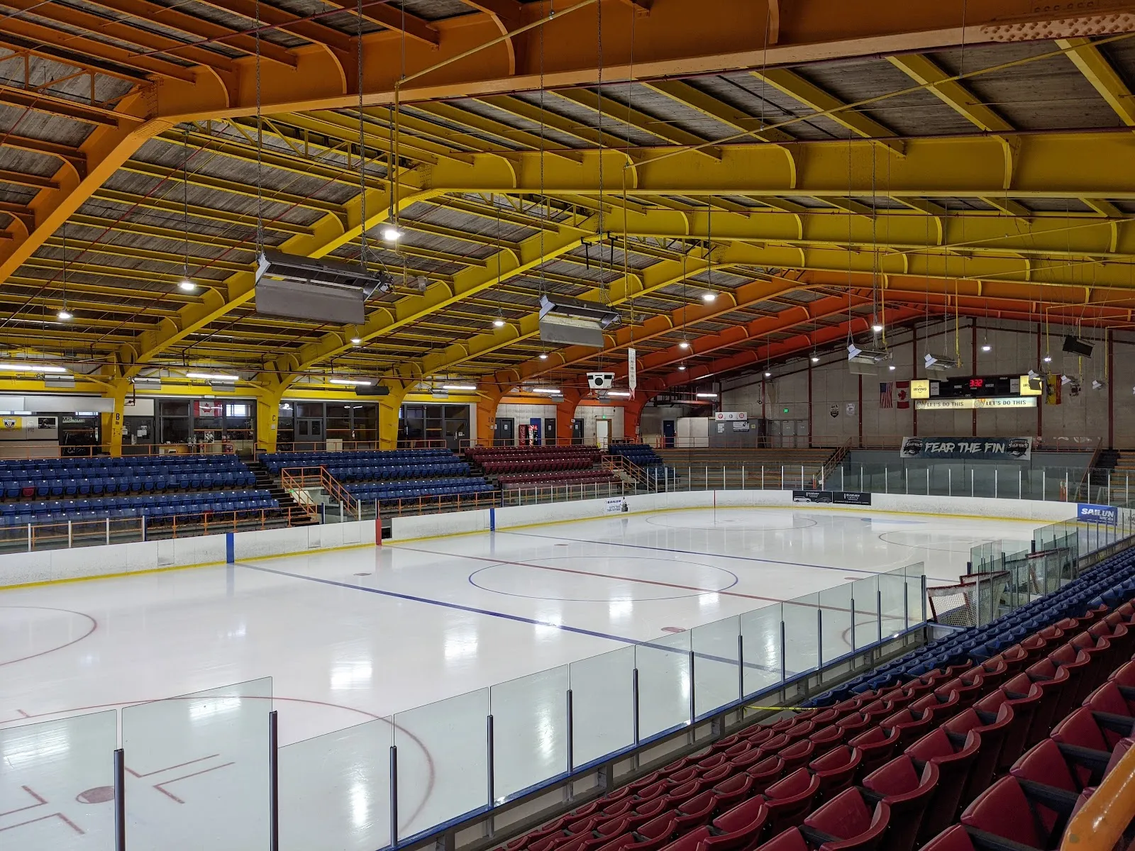 The empty ice seen from the red seats in the corner, under orange and yellow roof trusses, with blue seats along the far side. — Lord Beaverbrook Rink