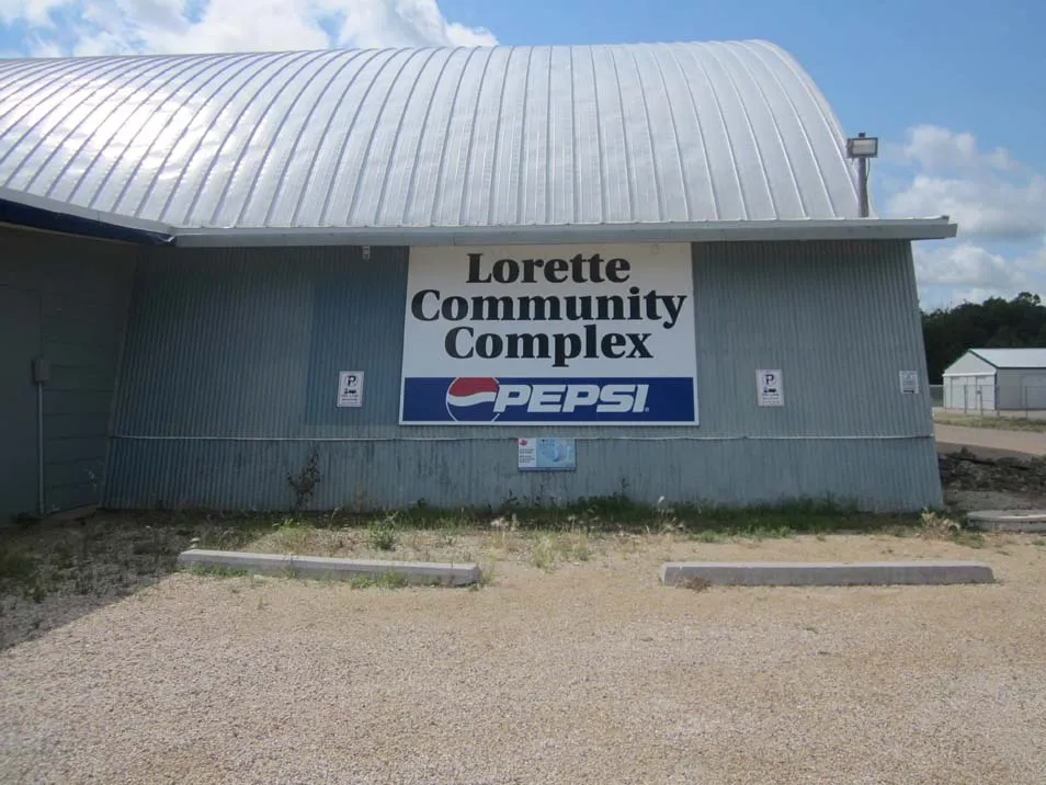 The end wall of the curved-roof arena building with a large 'Lorette Community Complex' sign, gravel parking in front. — Lorette Community Complex