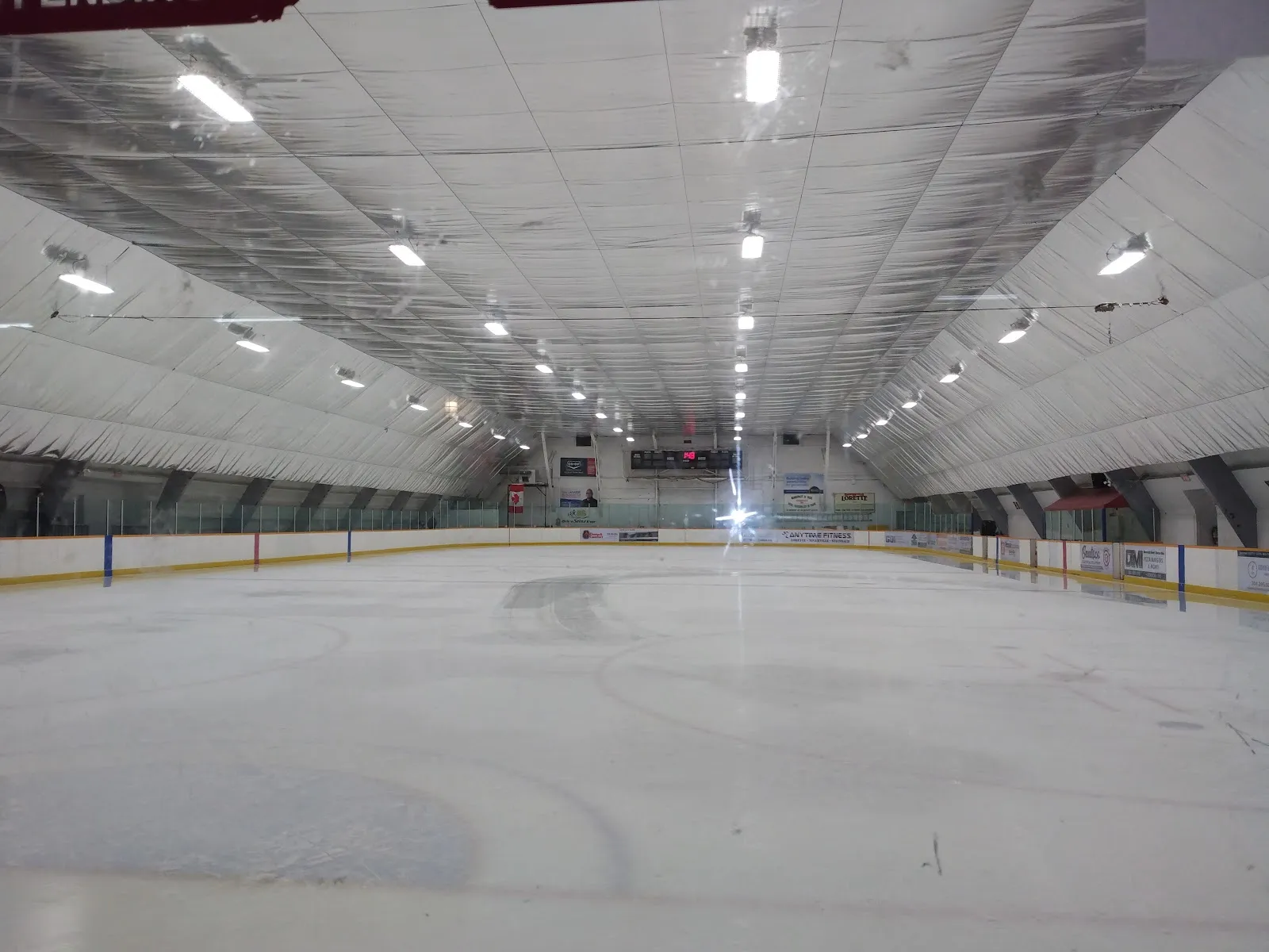 The empty ice from rink level, under a white insulated ceiling with rows of lights, boards running to the far end. — Lorette Community Complex