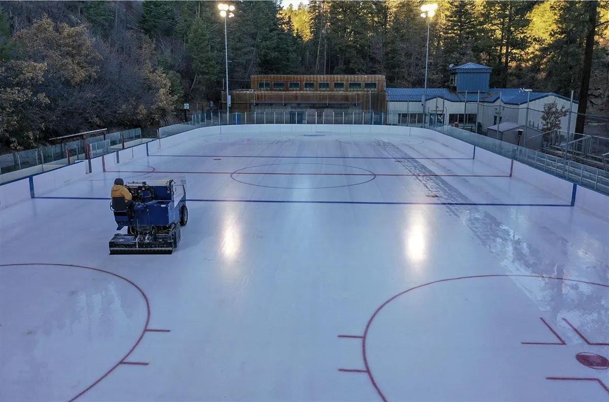 Aerial view of the Los Alamos County ice rink with a resurfacer on the clear ice and the outdoor rink's boards, trees and buildings around it. — Los Alamos County Ice Rink