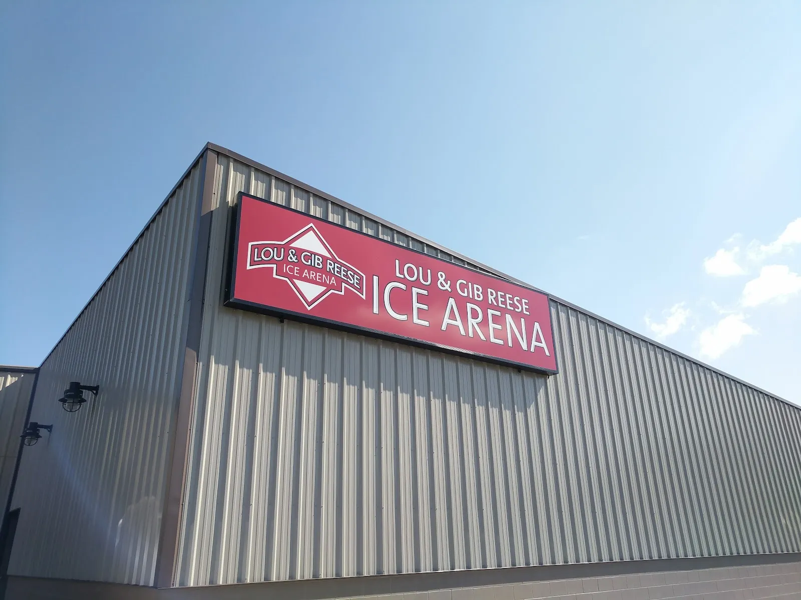 Corner of the metal-sided arena building, shot from below, with the red "Lou & Gib Reese Ice Arena" sign on the roofline against a clear sky — Lou & Gib Reese Ice Arena