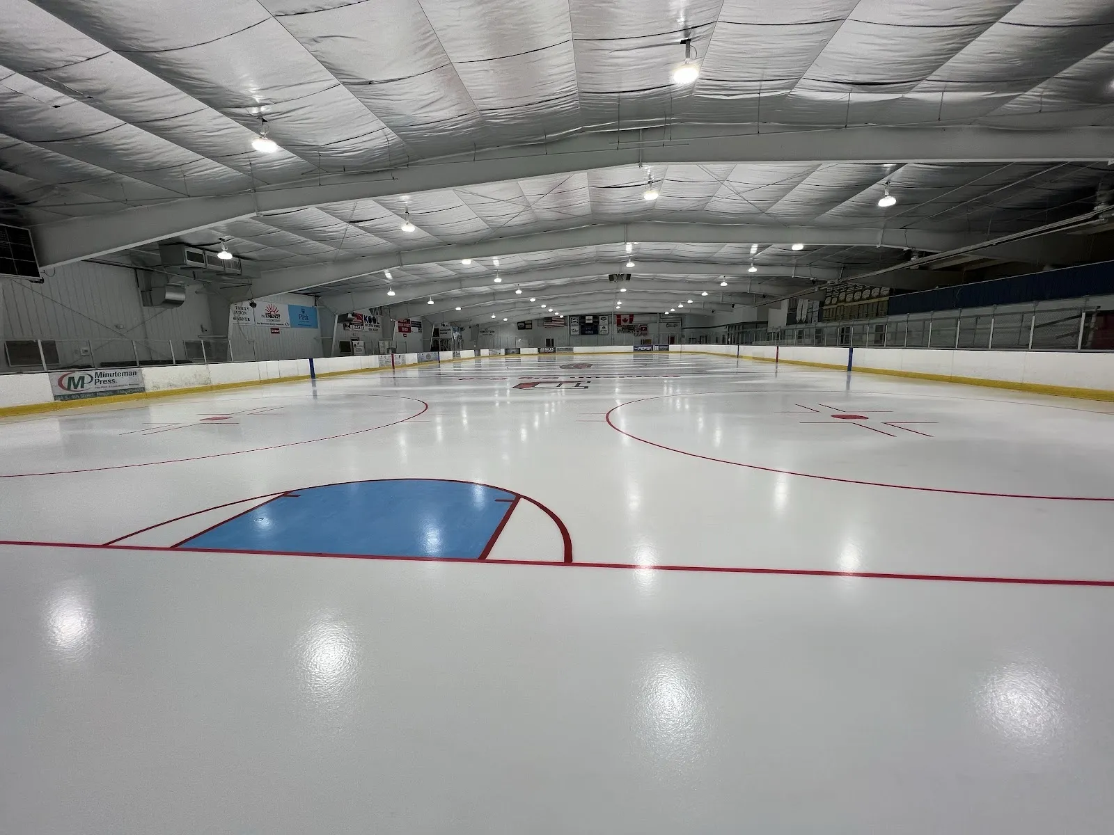 An empty sheet seen from the corner of the boards, a blue faceoff circle and red goal line on white ice under a pale curved ceiling. — Lou & Gib Reese Ice Arena