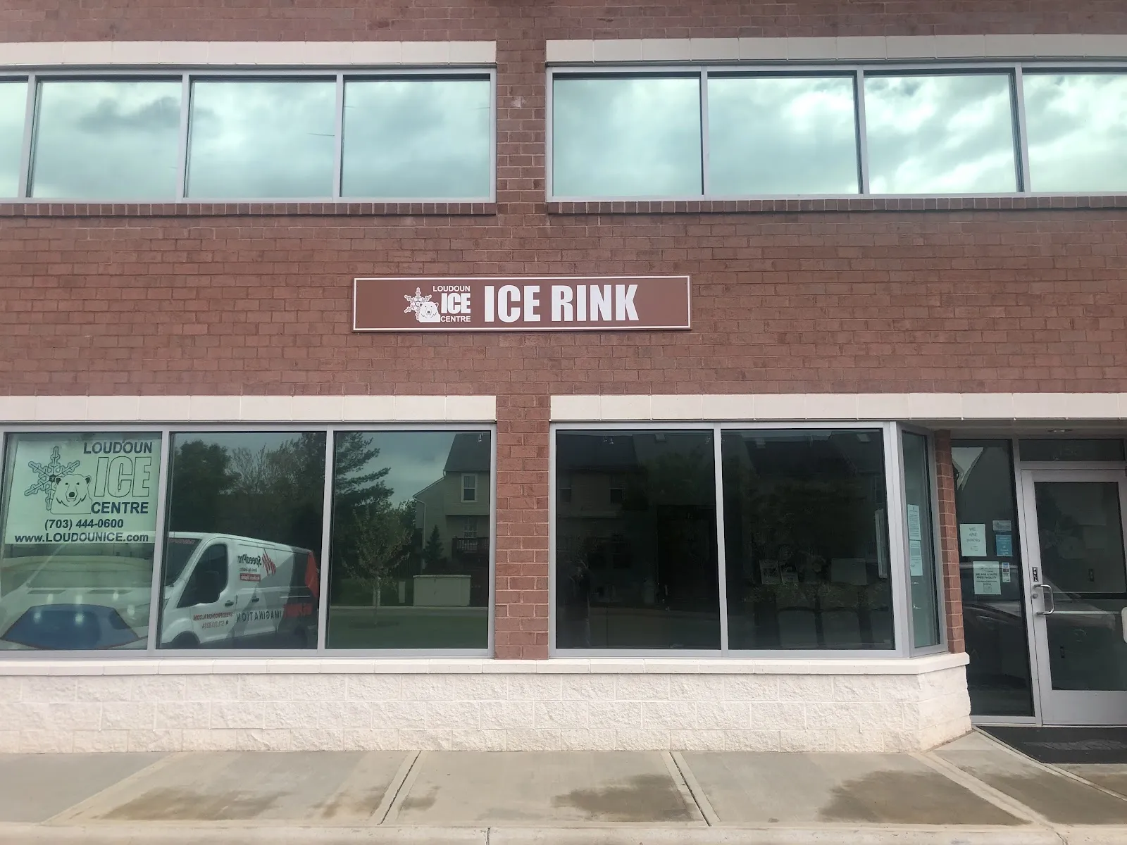 Fachada de local comercial de ladrillo con un letrero café de ICE RINK con el logotipo de Loudoun Ice Centre, ventanas en la planta baja, una de ellas rotulada LOUDOUN ICE CENTRE con una camioneta con la marca estacionada al frente, y una puerta de entrada de vidrio — Loudoun Ice Centre (Sterling)
