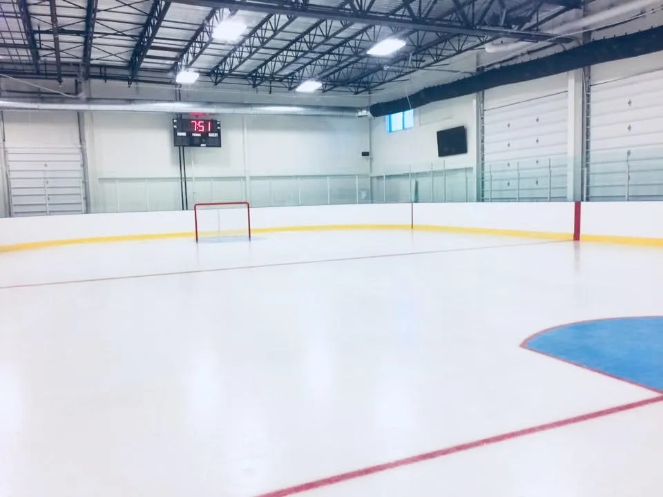Una pista vacía vista desde el extremo, con un círculo de saque azul y una línea roja sobre hielo blanco, con una portería en el extremo opuesto bajo un techo de armaduras de acero. — Loudoun Ice Centre (Sterling)