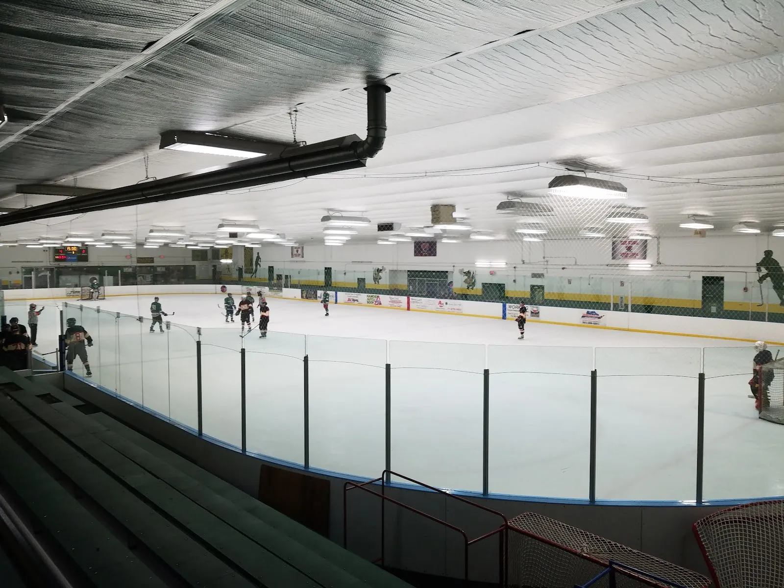 La pista vista desde las gradas detrás del vidrio, con patinadores repartidos por el hielo bajo un techo bajo y blanco. — Louis Astorino Ice Arena