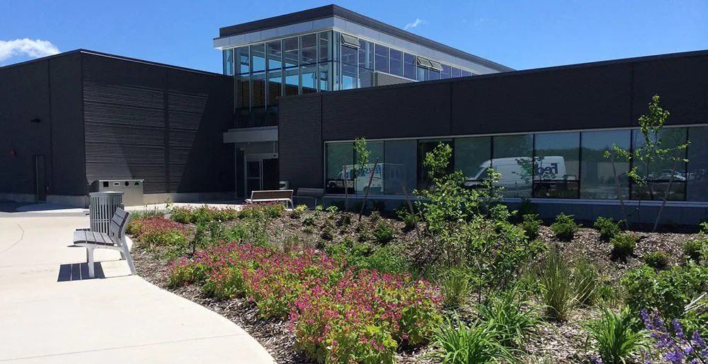 La fachada con revestimiento oscuro del centro, con un nivel superior de muros de vidrio y una plaza ajardinada con arbustos en flor y bancas bajo el sol de verano. — Lunenburg County Lifestyle Centre