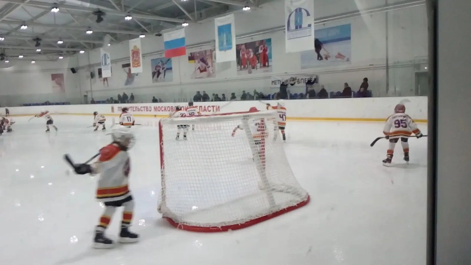 Children playing hockey in front of a goal on the Ramenskoye ice sheet, with club banners above the boards — Lutchenko Ice Arena Ramenskoye