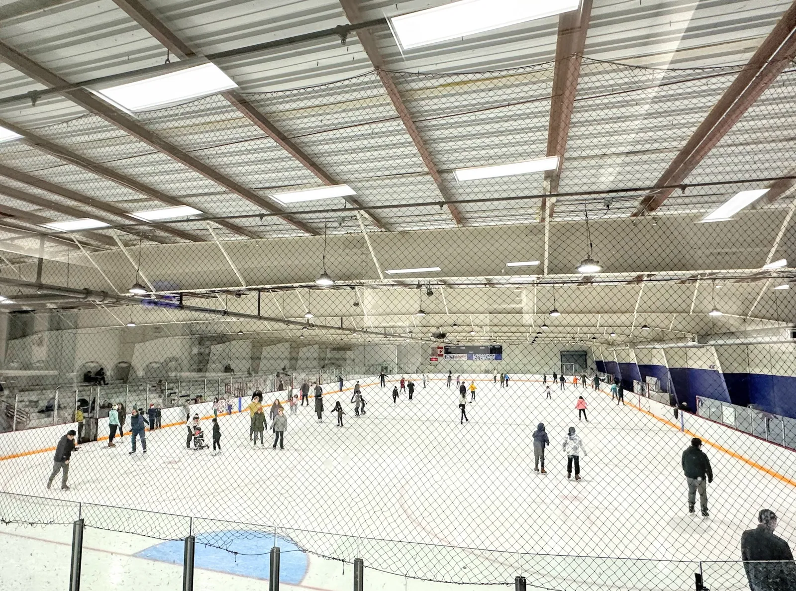 A public skating session seen from the end of the rink through the netting, under a white roof with timber beams and banners along the far wall. — Lynnwood Ice Center