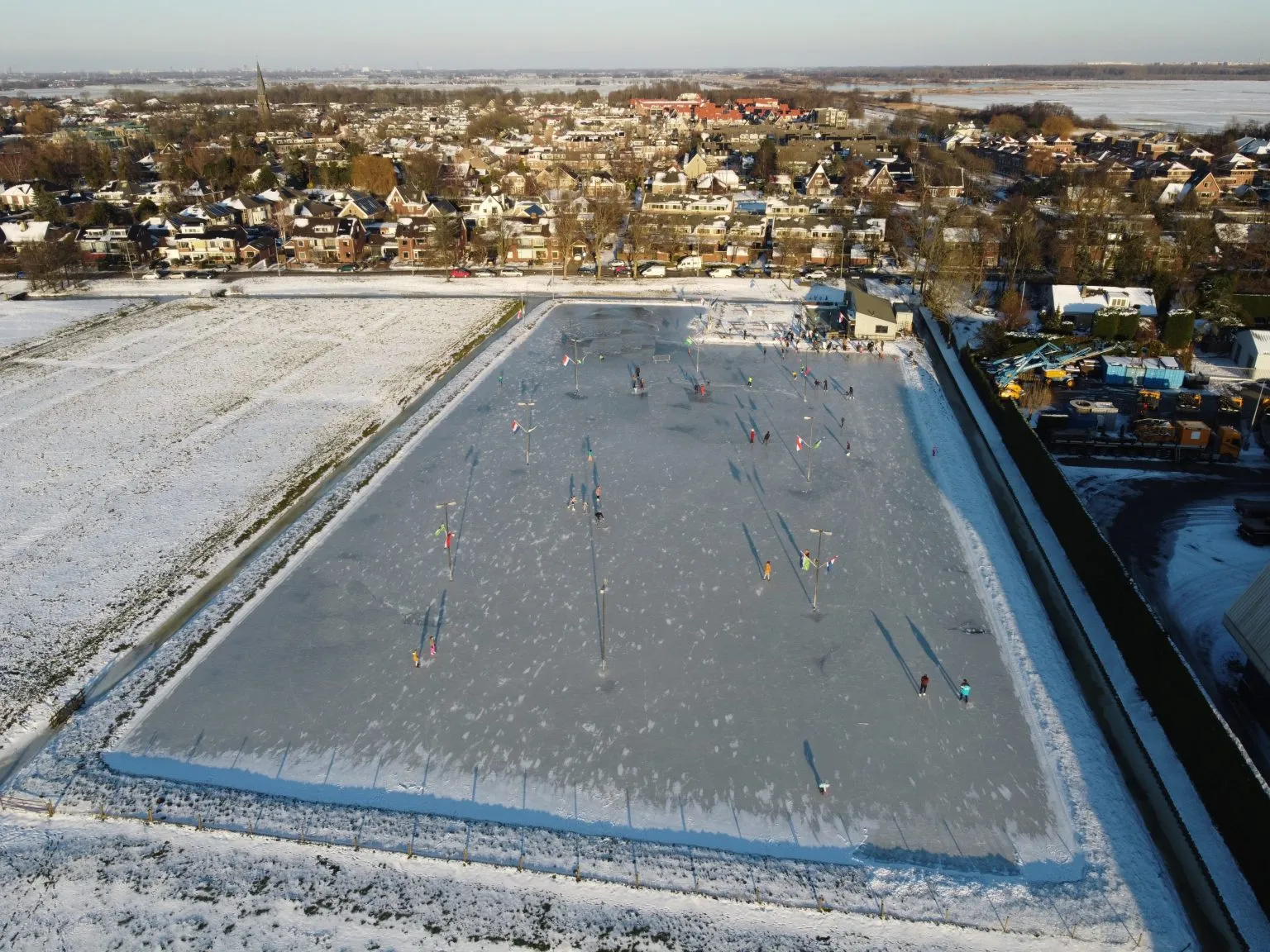 An aerial view of the swept natural-ice rink at Maasland in February 2021 with skaters, flag-topped lamp posts and the clubhouse at the far end, snowy fields on one side and the village behind. — Landijsbaan Maasland (Maaslands IJsvermaak)