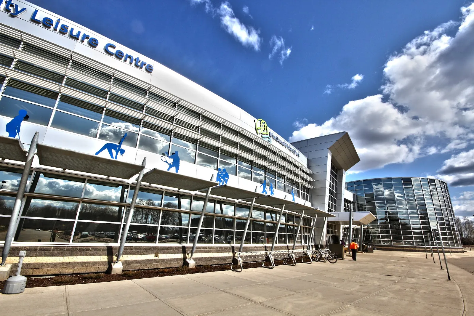 The glass-and-steel front of the Suncor Community Leisure Centre at MacDonald Island Park under a bright sky, seen from the walkway, with the curved wing beyond. — MacDonald Island Park