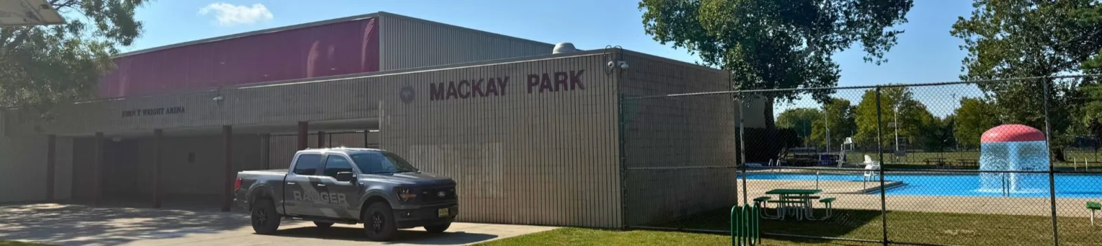 Vista de día del edificio de John T. Wright Arena con revestimiento metálico color arena en Mackay Park, con letreros de "MACKAY PARK" y "JOHN T WRIGHT ARENA" en la pared y la alberca al aire libre contigua visible detrás de una cerca de malla ciclónica. — Mackay Ice Rink