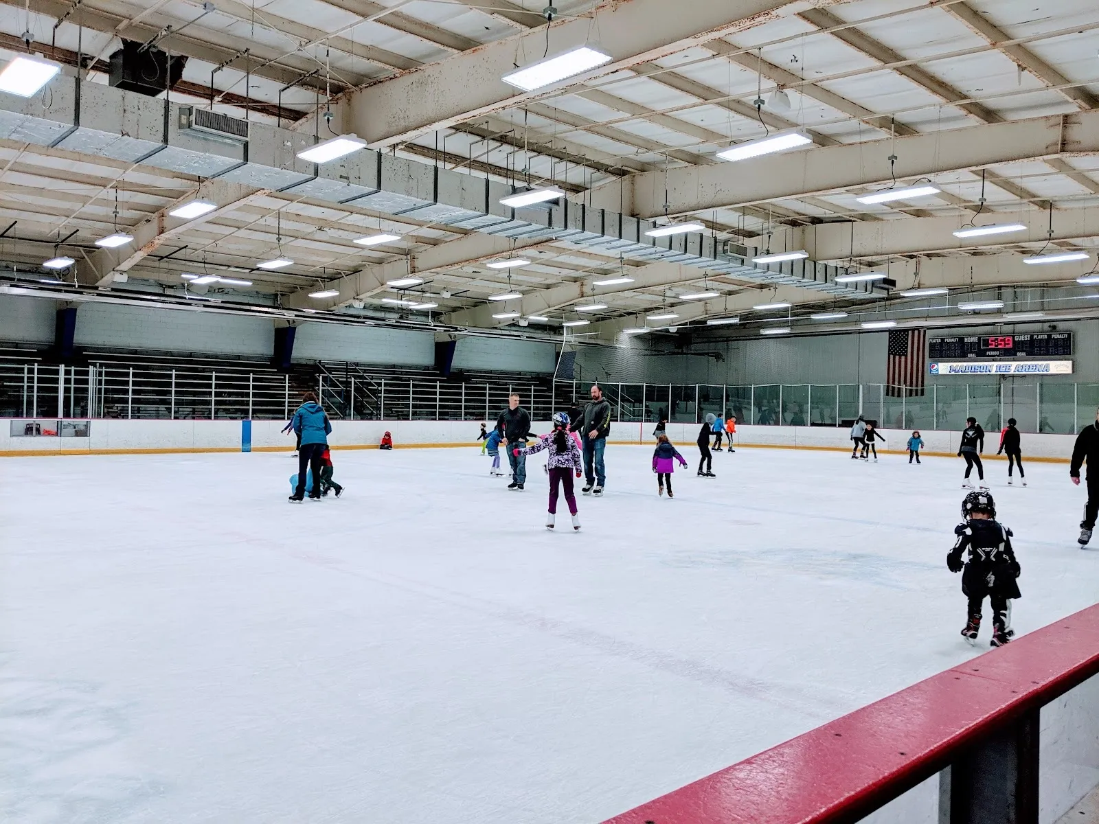Una sesión pública en una pista amplia, con patinadores repartidos por el hielo detrás de un barandal de remate rojo bajo un largo techo de luces. — Madison Ice Arena