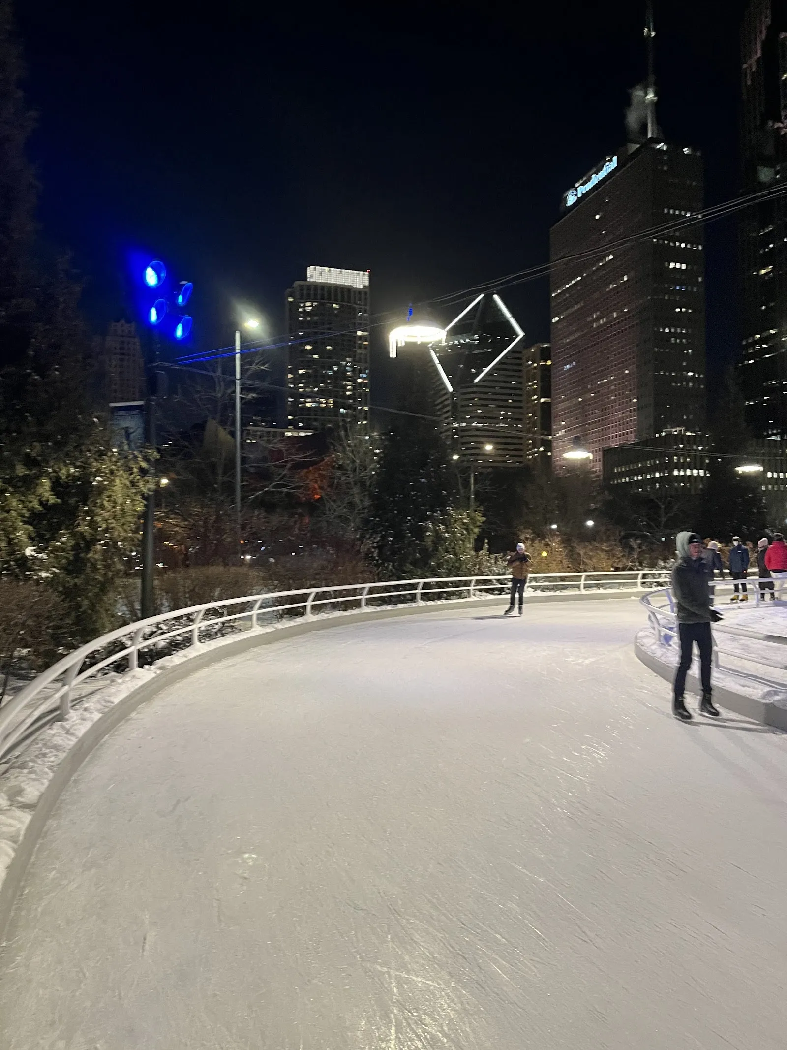 El Skating Ribbon serpenteando por Maggie Daley Park, con unos cuantos patinadores en el hielo y, detrás, el muro de escalada y los rascacielos de Chicago bajo un cielo azul. — Maggie Daley Park Ice Skating Ribbon
