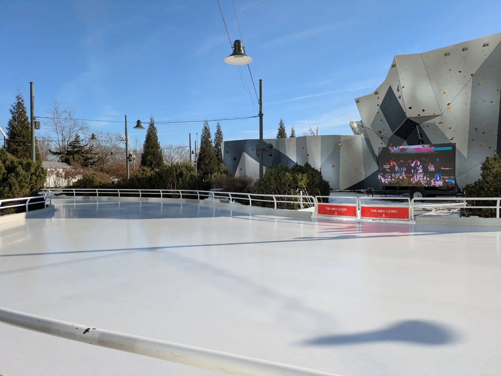 Un tramo vacío y recién pulido de la cinta en un día soleado, con su barandal blanco curvándose hacia las coníferas y el muro de escalada facetado. — Maggie Daley Park Ice Skating Ribbon