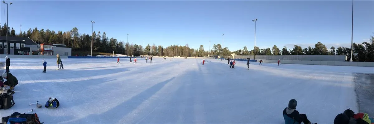 Wide view of the ice at the Maier Arena with skaters in the distance and skating bags along the near edge. — Maier Arena