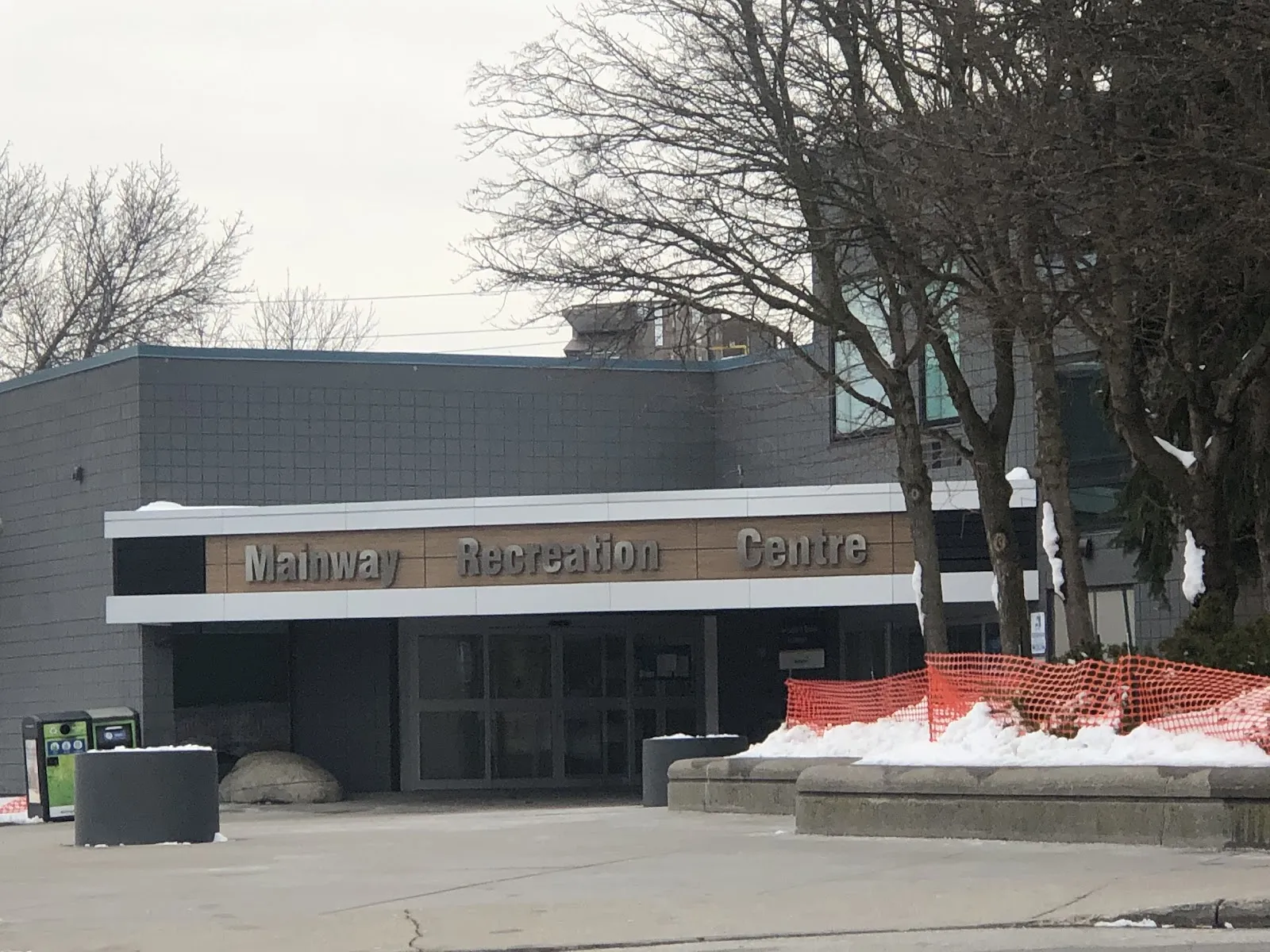 Front entrance of Mainway Recreation Centre on a winter day, grey tiled facade with a wood-toned sign reading "Mainway Recreation Centre" above the glass doors, bare trees and orange snow fencing in front. — Mainway Ice Centre (Mainway Recreation Centre)
