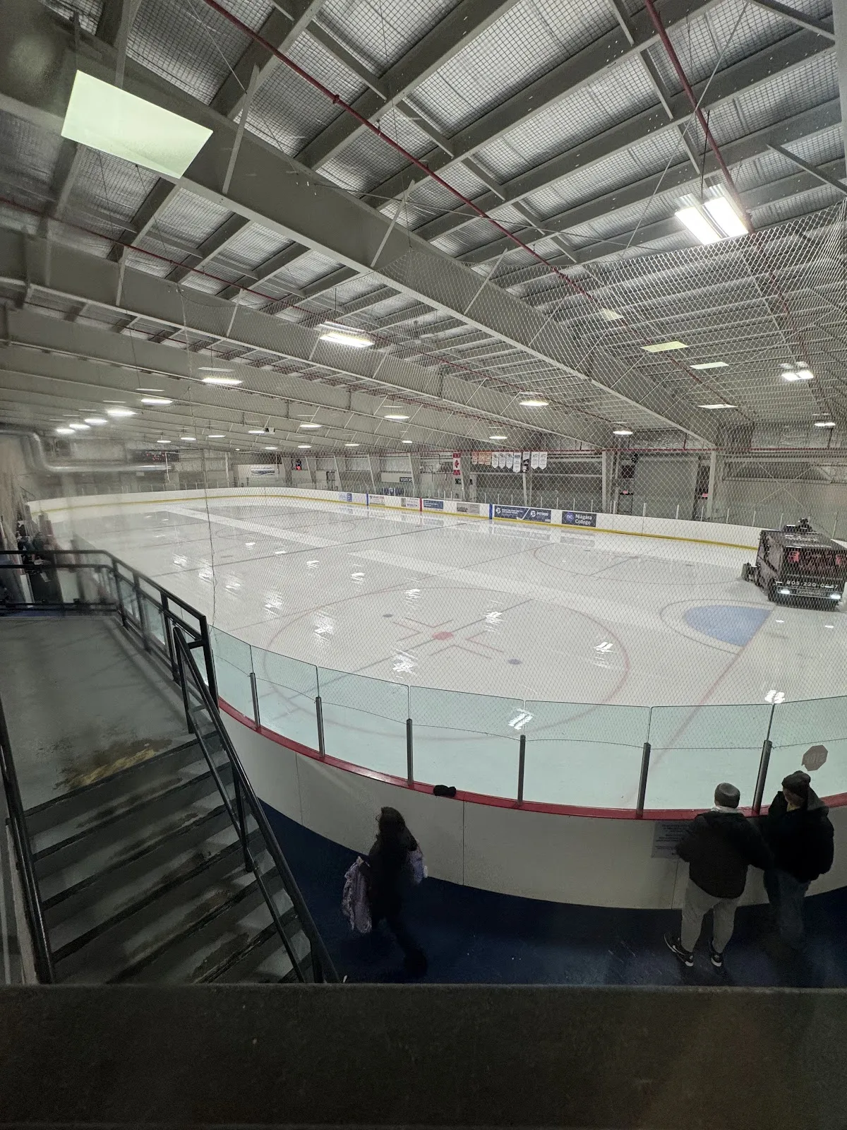 Elevated view of an empty ice sheet at Mainway Ice Centre from the upper walkway, boards and glass along the near side, a Zamboni parked on the ice, sponsor ad boards along the far boards. — Mainway Ice Centre (Mainway Recreation Centre)