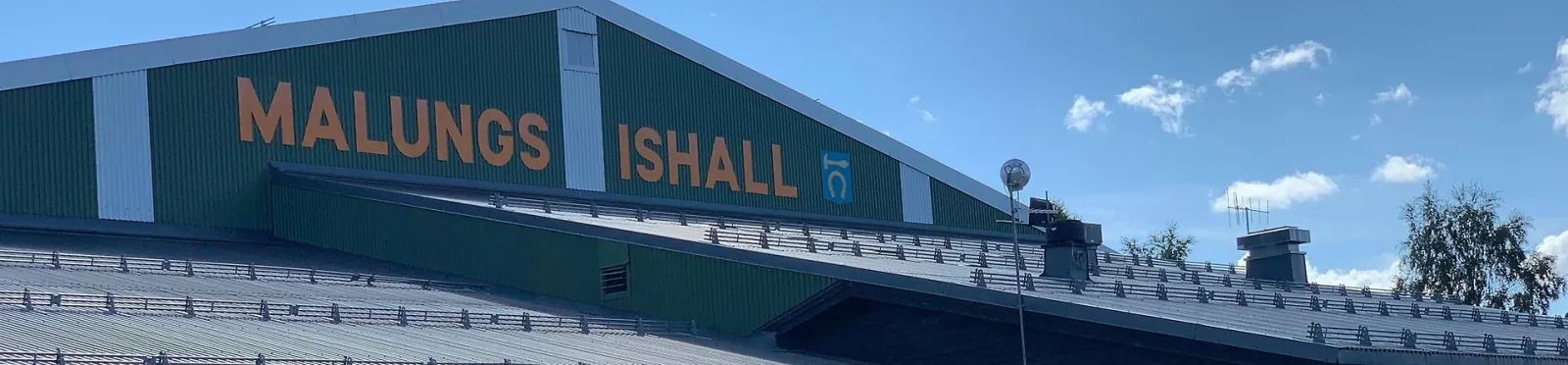 The roofline of the Malung ice hall with its green wall and the lettering MALUNGS ISHALL, seen from outside against a blue sky. – Malungs ishall