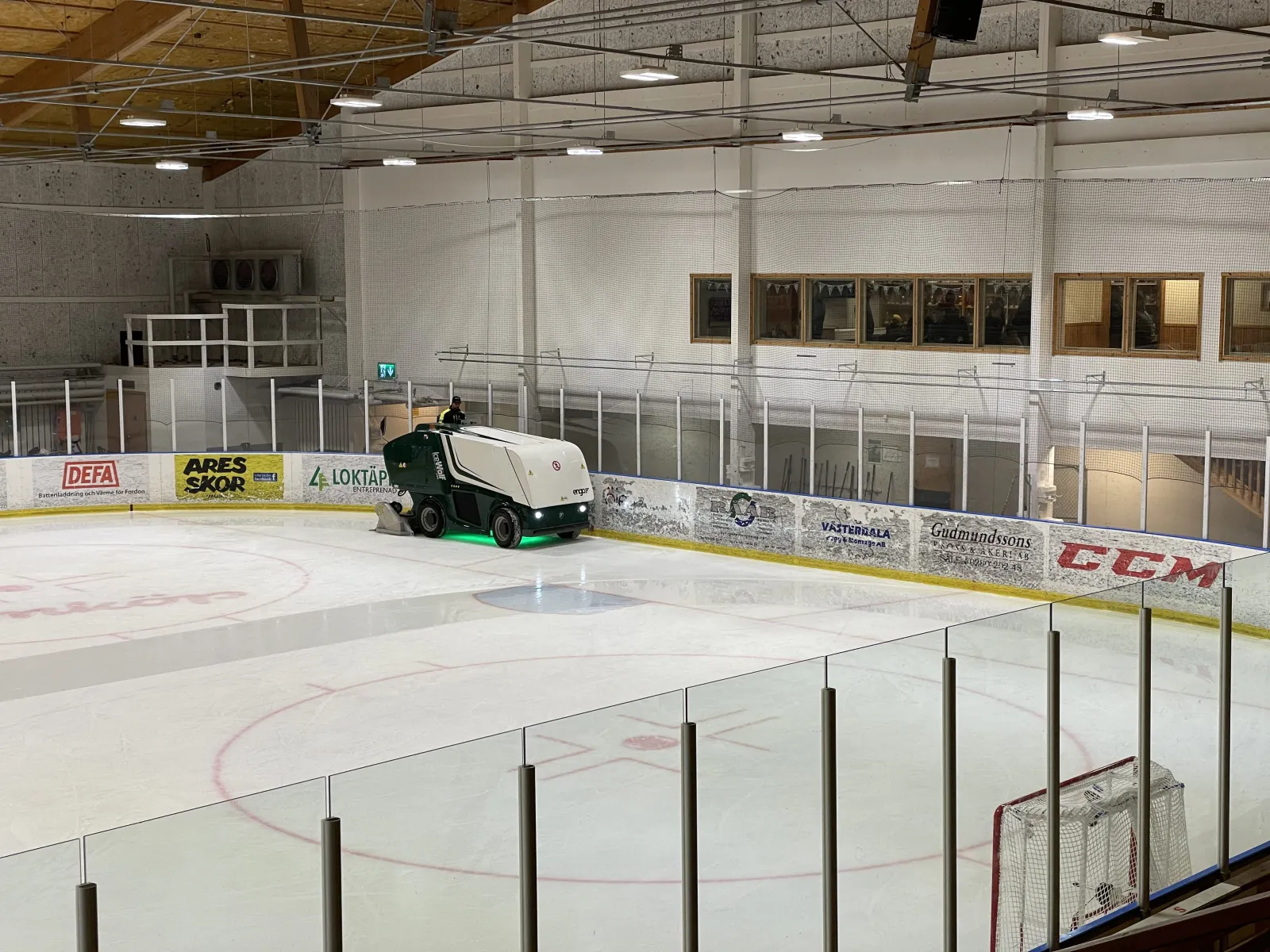 The ice sheet of Malungs ishall with the ice-resurfacing machine at the far boards, glass-topped boards, sponsor panels and a timber-beamed roof above. – Malungs ishall