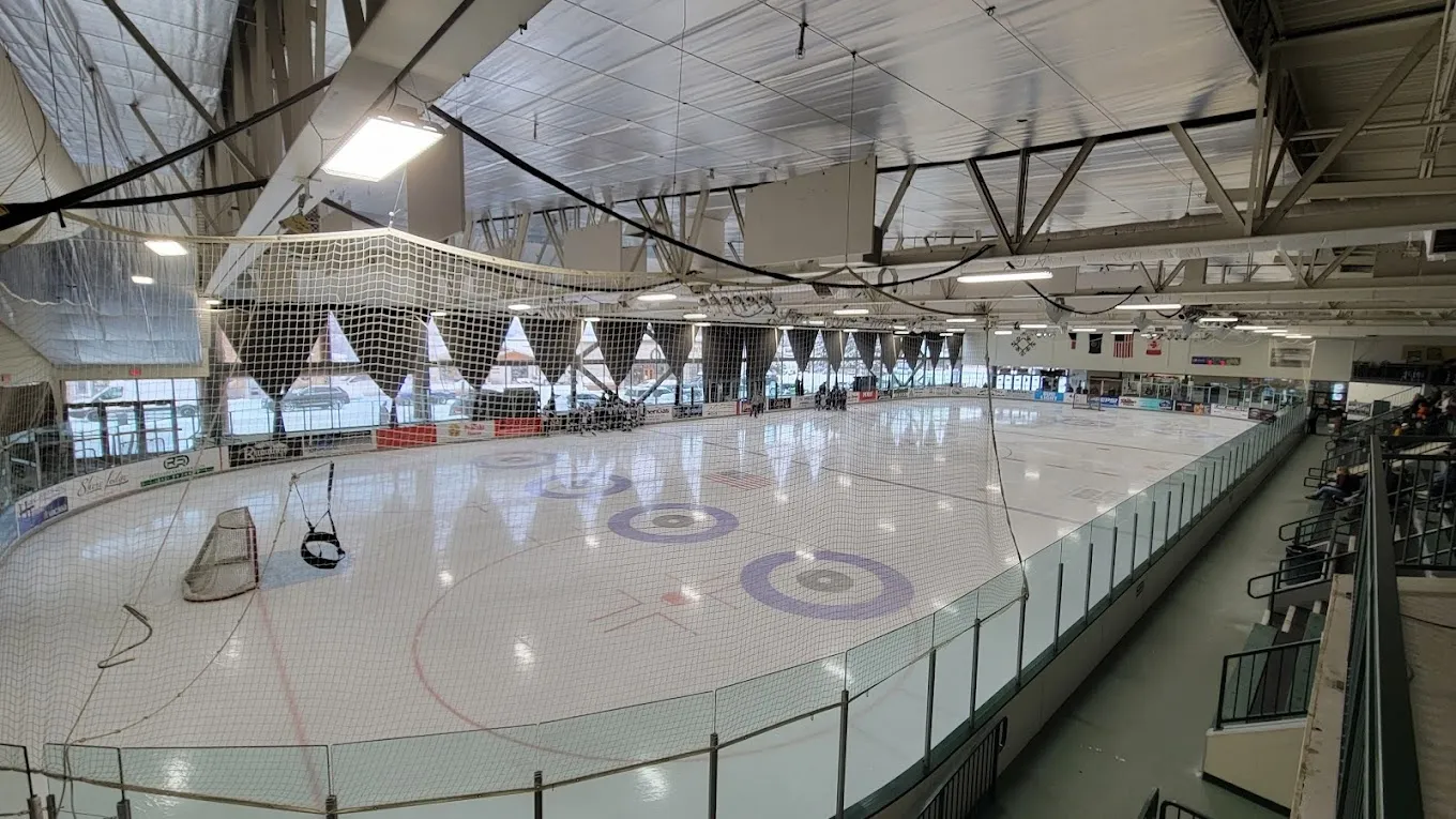 A wide view of the sheet through a glass post, the empty ice with a red-ringed centre circle under a steel-trussed roof with big windows on the far wall. — Manchester Ice & Events Centre