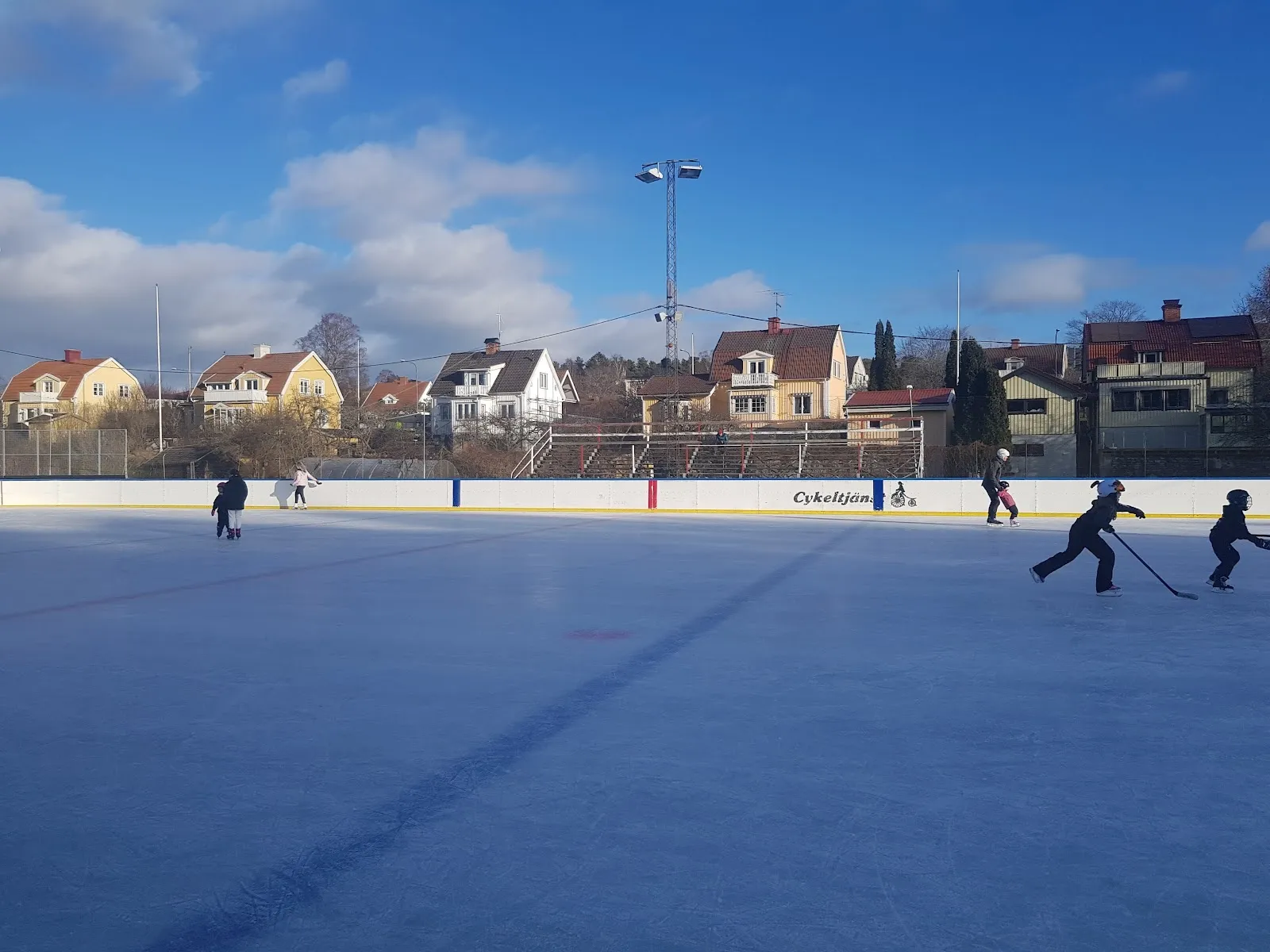 Utomhusrinken Månskensrinken i Södertälje i skymningen, med åkare på det blåbelysta isen och husen och strålkastarna bortom. – Månskensrinken