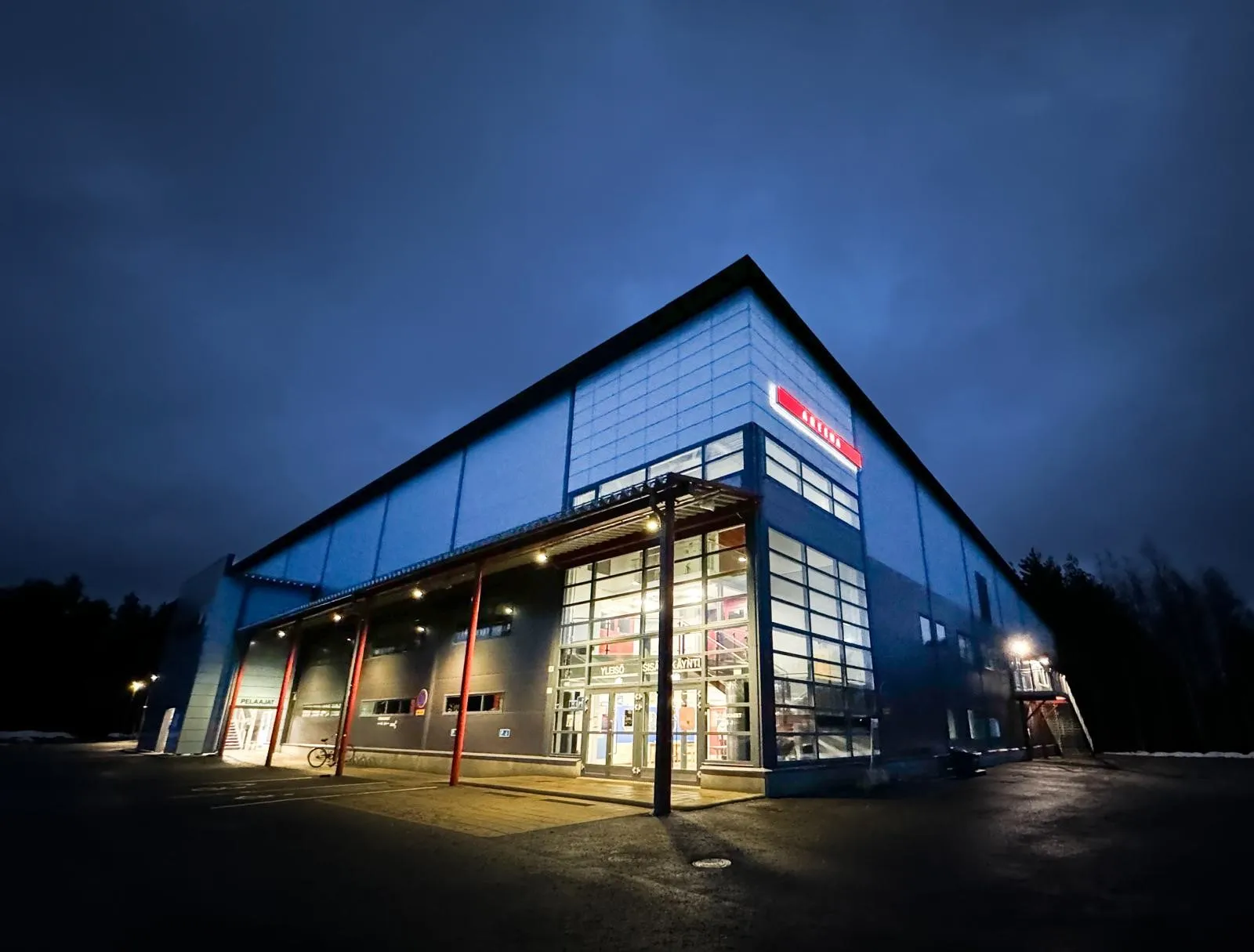 The blue and grey Mäntän Jääareena building at dusk with its lit glass entrance canopy and red supports, seen from the car park. — Mäntän Jääareena