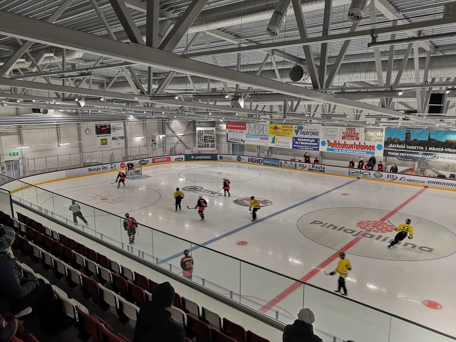 A wide view of the Mänttä ice arena from the stand, with players spread across the ice and the boards beyond. — Mäntän Jääareena