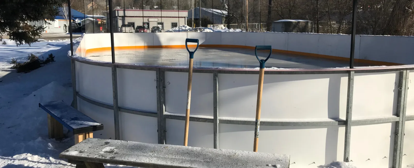 La pequeña pista con vallas de Maple Avenue en invierno, con palas para raspar el hielo apoyadas en las vallas y un banco al lado. — Maple Avenue Rink (Haliburton)
