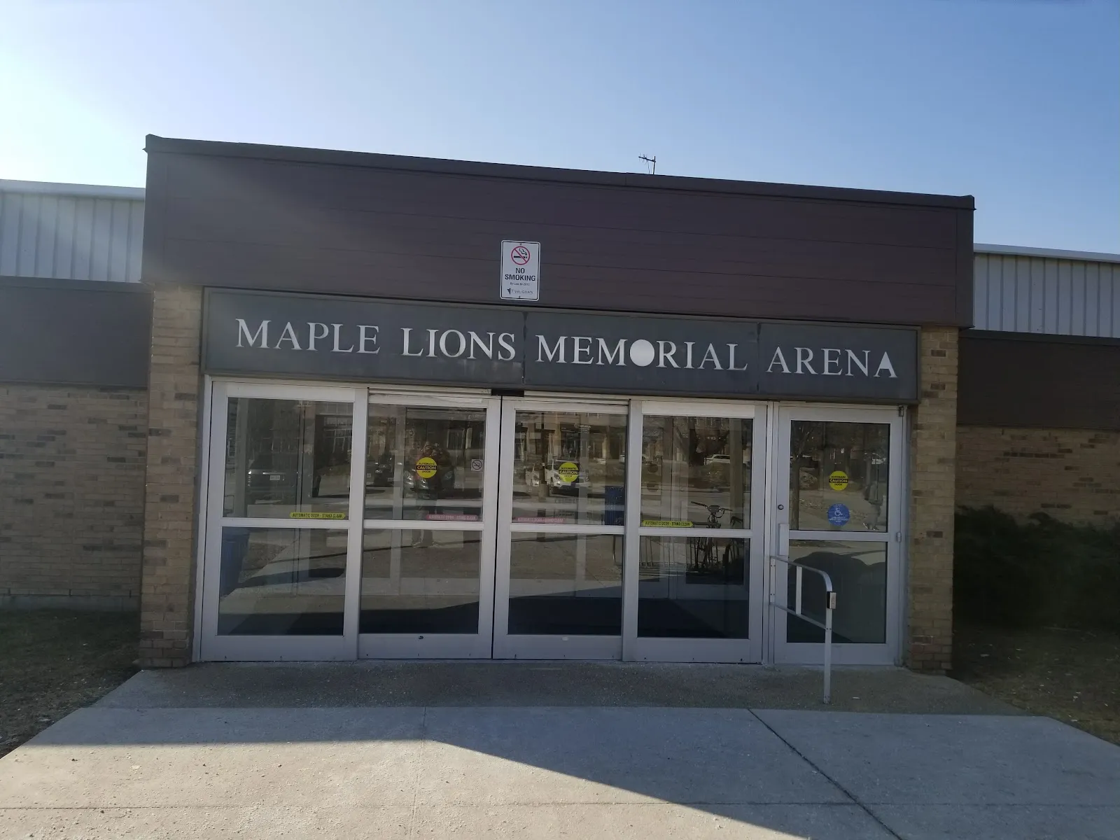 The entrance to the Maple Lions Memorial Arena at the Maple Community Centre: glass double doors under the arena name on a brick facade, with a paved forecourt and clear sky. — Maple Community Centre (Vaughan)