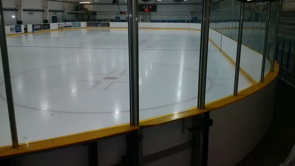 The ice from the boards behind the glass: the centre line and faceoff circle with the far end and scoreboard beyond. — Maple Community Centre (Vaughan)