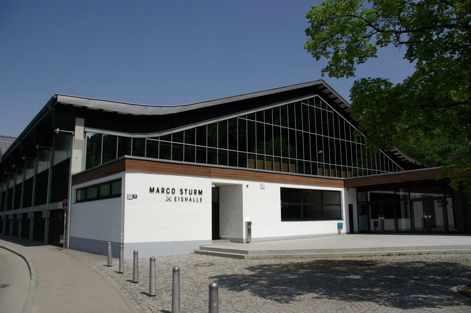 Das weiße Eingangsgebäude mit dem Schriftzug Marco Sturm Eishalle und dahinter das verglaste, wellenförmige Dach der Halle. – Marco-Sturm-Eishalle