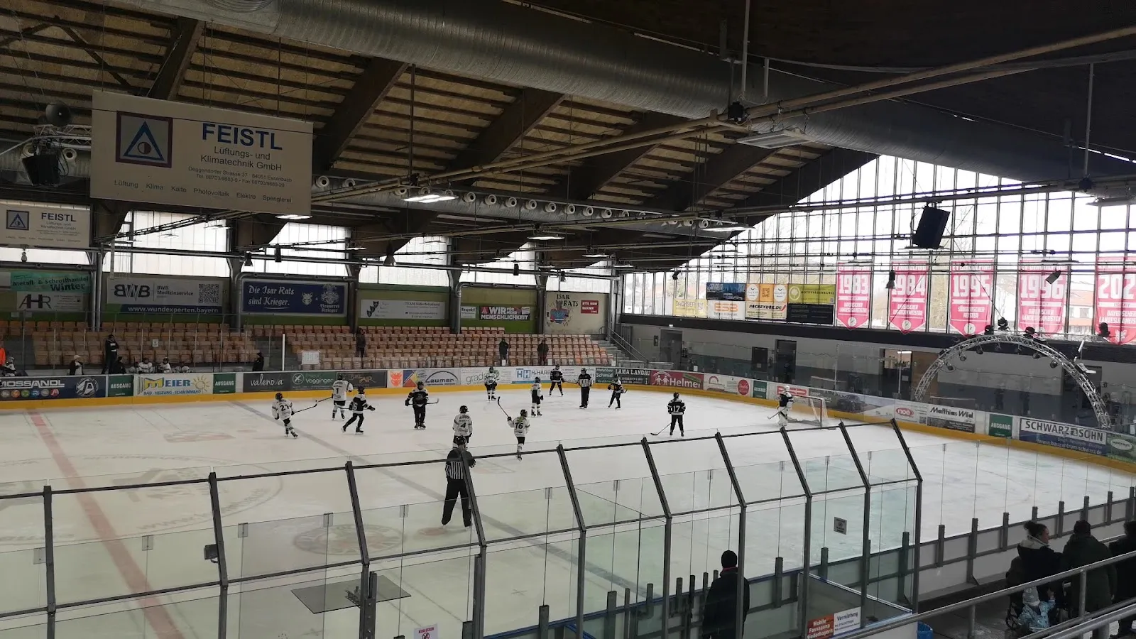Ein Nachwuchs-Eishockeyspiel auf der einzigen Eisfläche der Halle, von der Tribüne unter dem Holzbinderdach aus gesehen, mit Schiedsrichtern und Spielern auf der ganzen Fläche. – Marco-Sturm-Eishalle