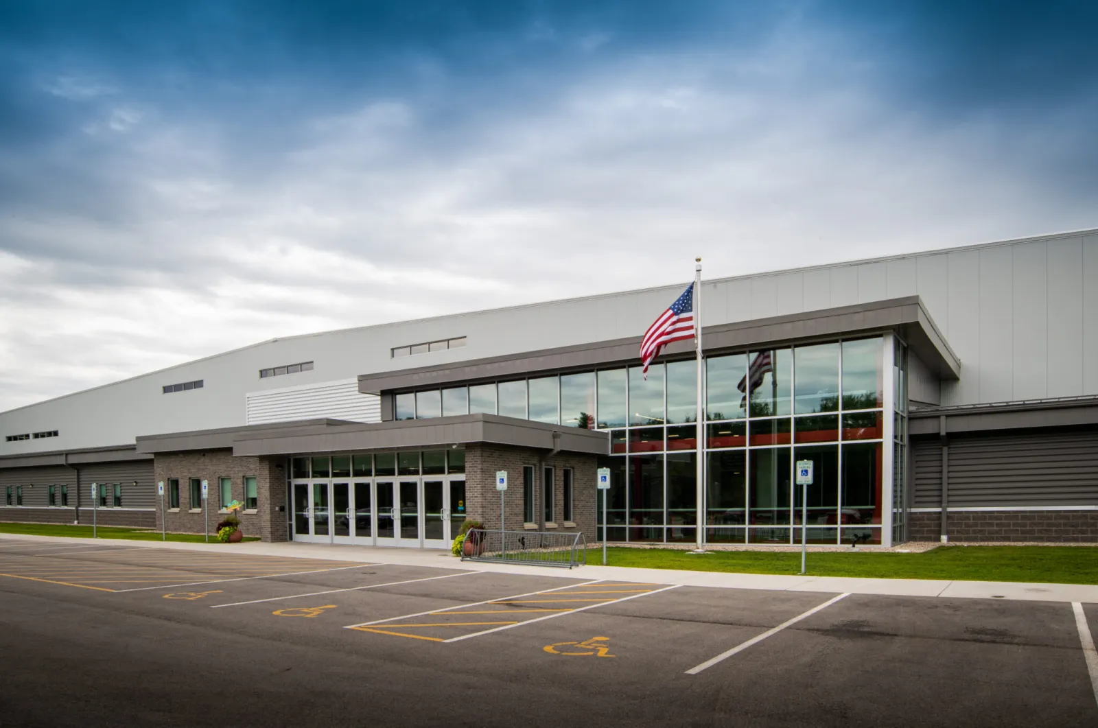 Front entrance of the Community REC Center, a large metal-panel building with a glass lobby, brick entry vestibule, flagpole, and handicap-marked parking lot in the foreground — Marinette Recreation Center