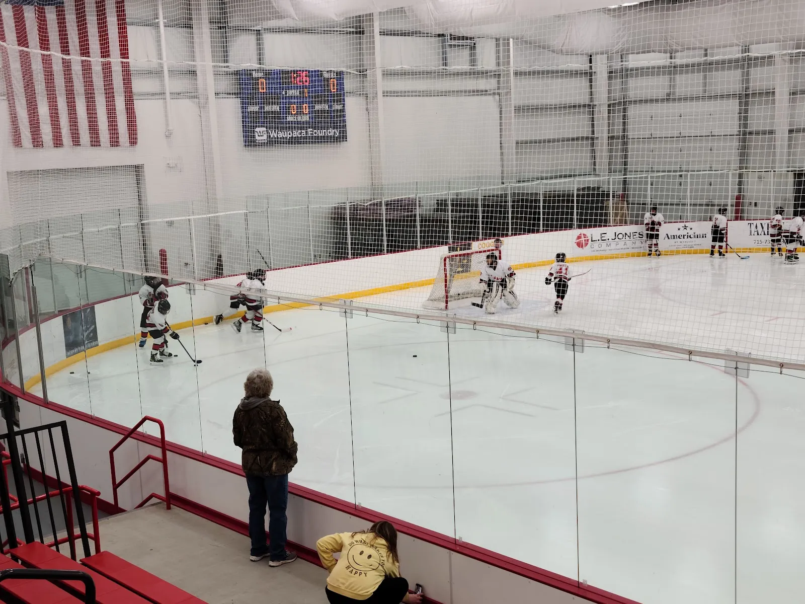 An ice sheet seen from the glass at the stands, with a youth game in play at the far end, the goal and a scoreboard behind it, and a few spectators along the boards in a bright steel-framed hall. — Marinette Recreation Center